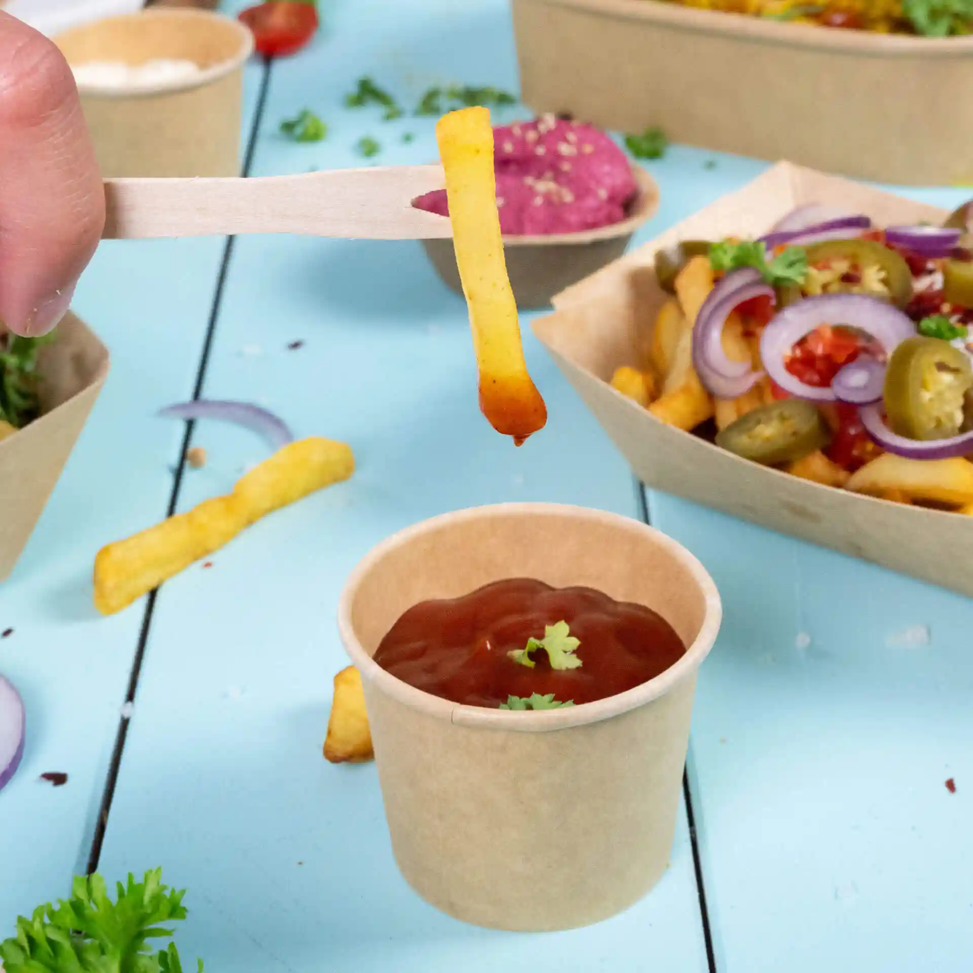 Bild einer Hand, die eine Holzgabel mit einer Pommes über einem kleinen Becher Ketchup auf einer blauen Oberfläche hält.