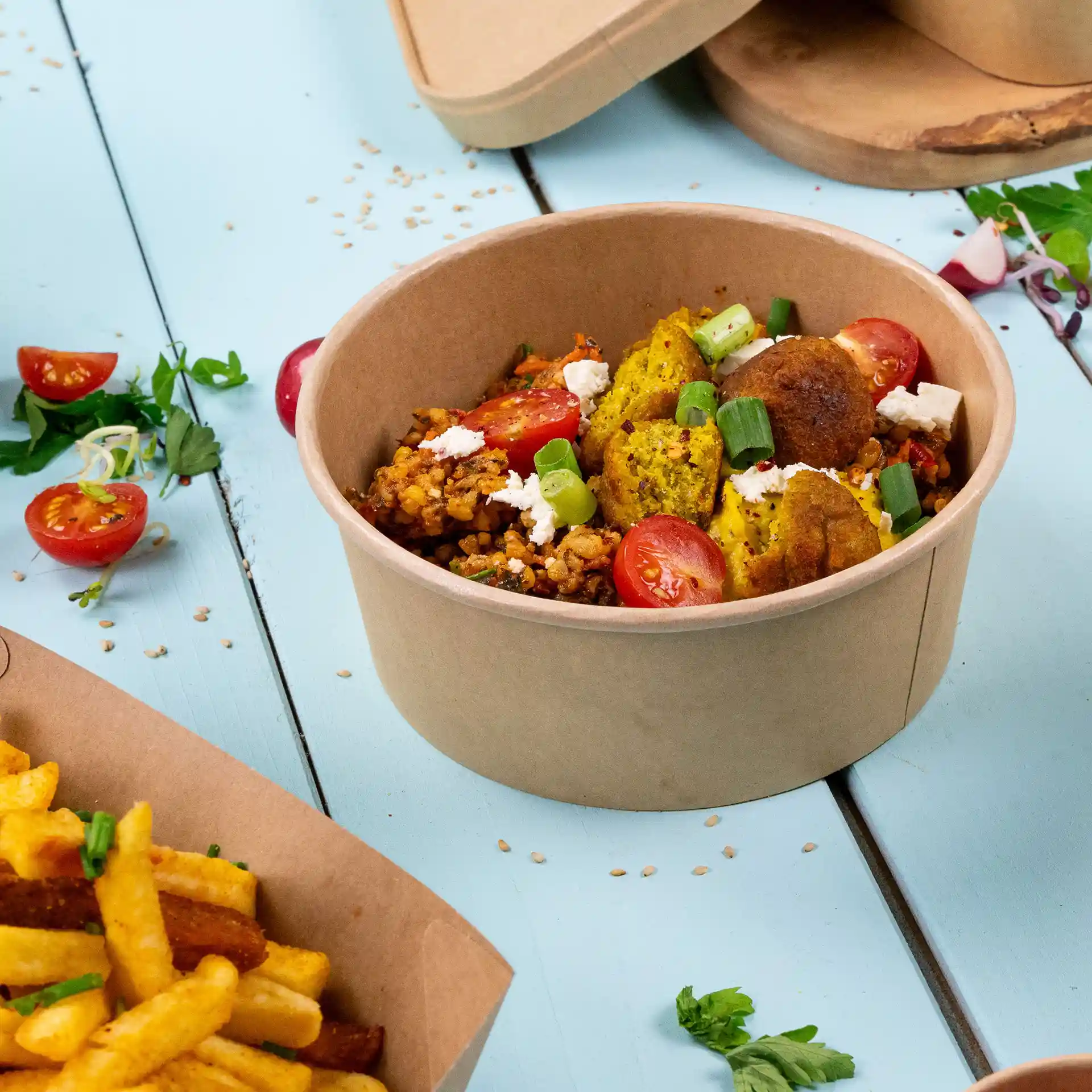Round brown paper bowl filled with grains, vegetables, and garnished with cherry tomatoes and green onions.