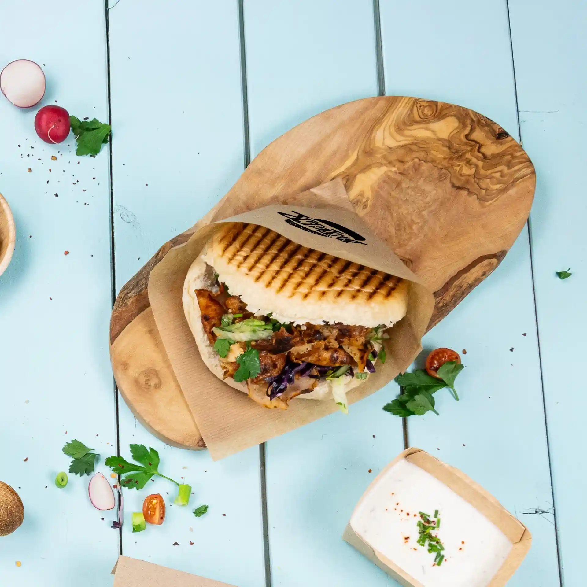Image of a grilled sandwich in a paper sleeve on a wooden board, with fresh herbs and a small container of sauce nearby.