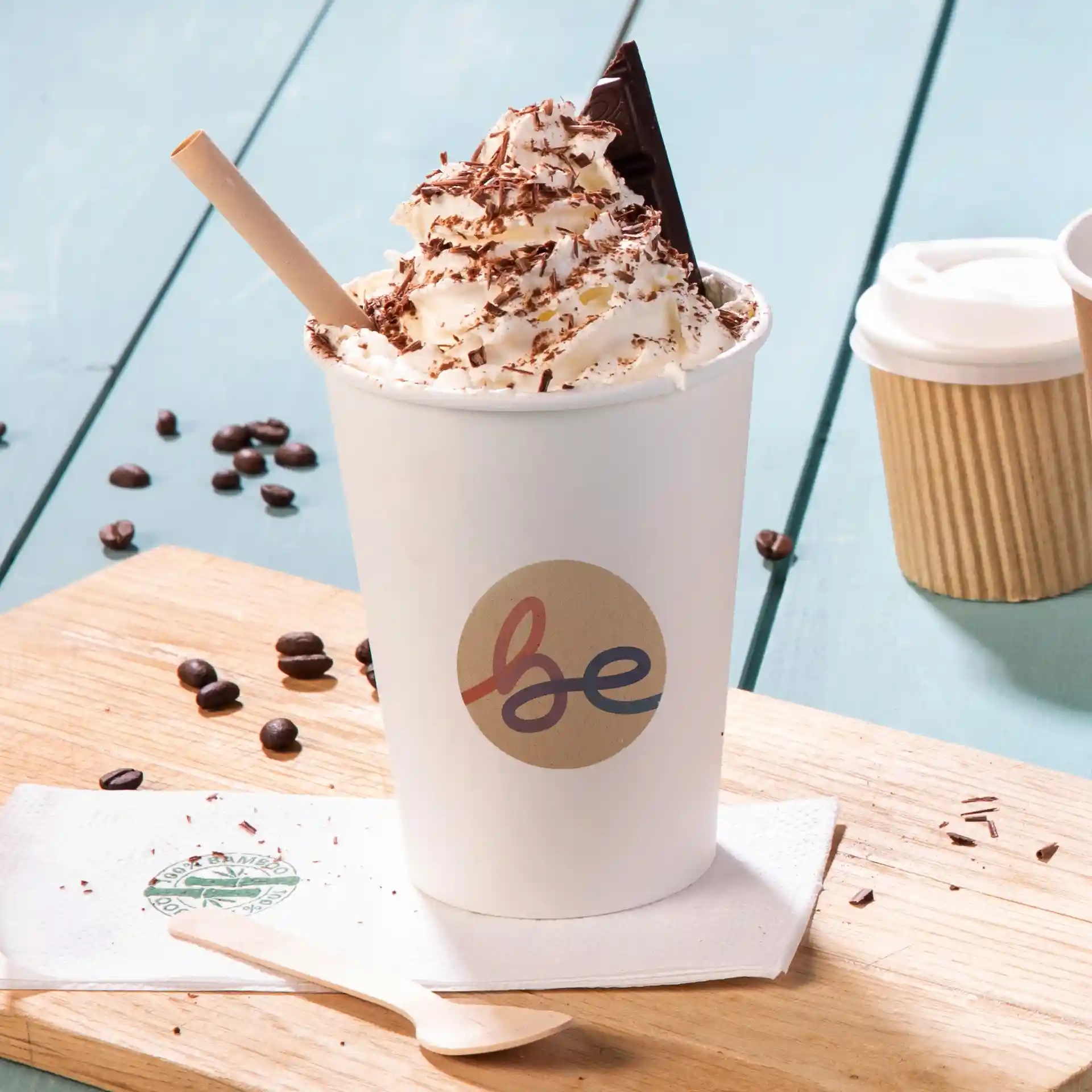 Cup with whipped cream, chocolate shavings, and a chocolate piece, on a wooden surface with coffee beans and a napkin.