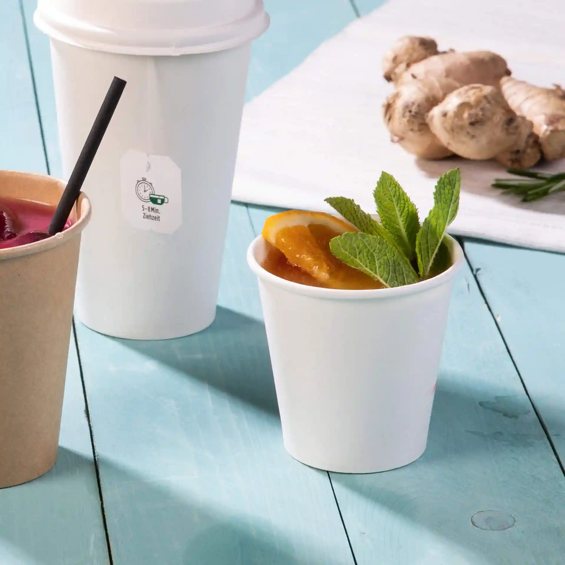 White paper cup with mint leaves and orange slices, placed on a blue wooden surface, with other cups in the background.