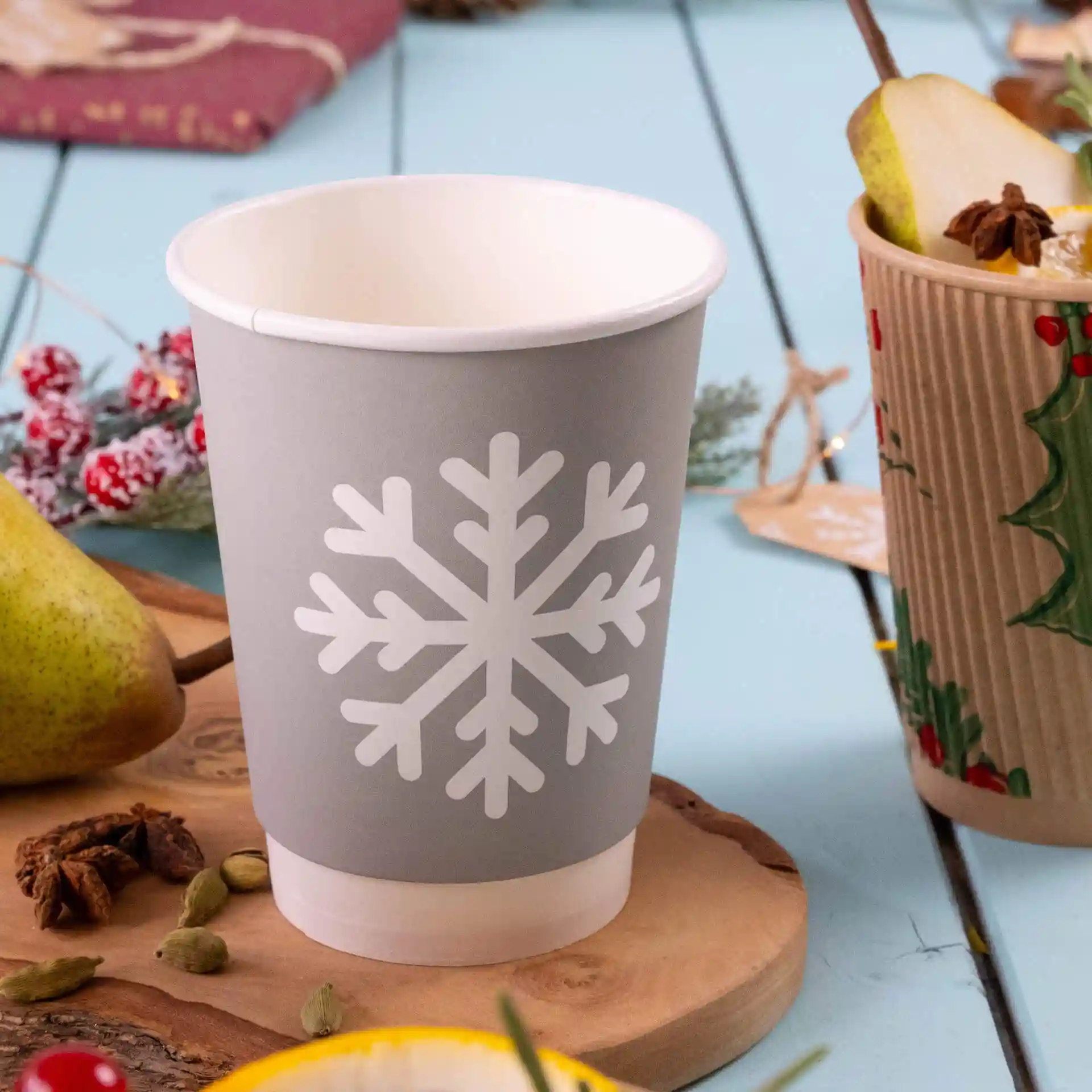 Grauer Becher mit weißem Schneeflocken-Design, doppelwandig, auf einer Holzoberfläche mit einer Birne und Gewürzen.