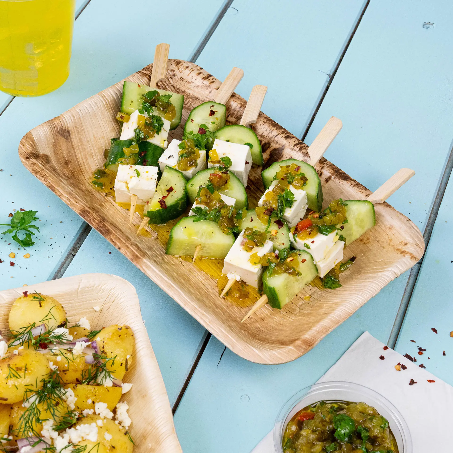 Ein rechteckiger Teller aus Palmblättern mit Gurken- und Feta-Spießen, garniert mit grüner Sauce und Koriander.