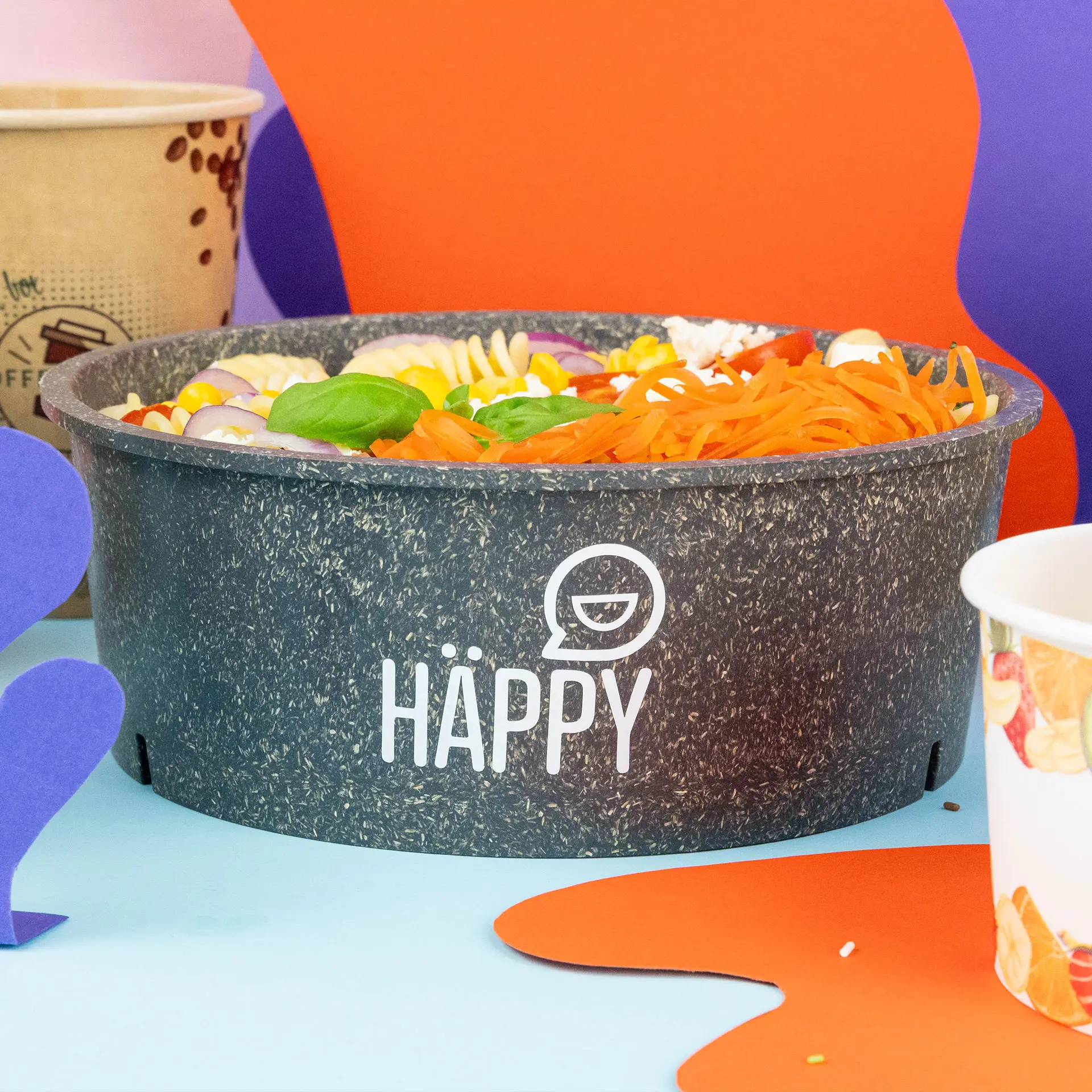 Reusable grey bowl with printed branding, containing colorful pasta and vegetables, placed on a light blue background.