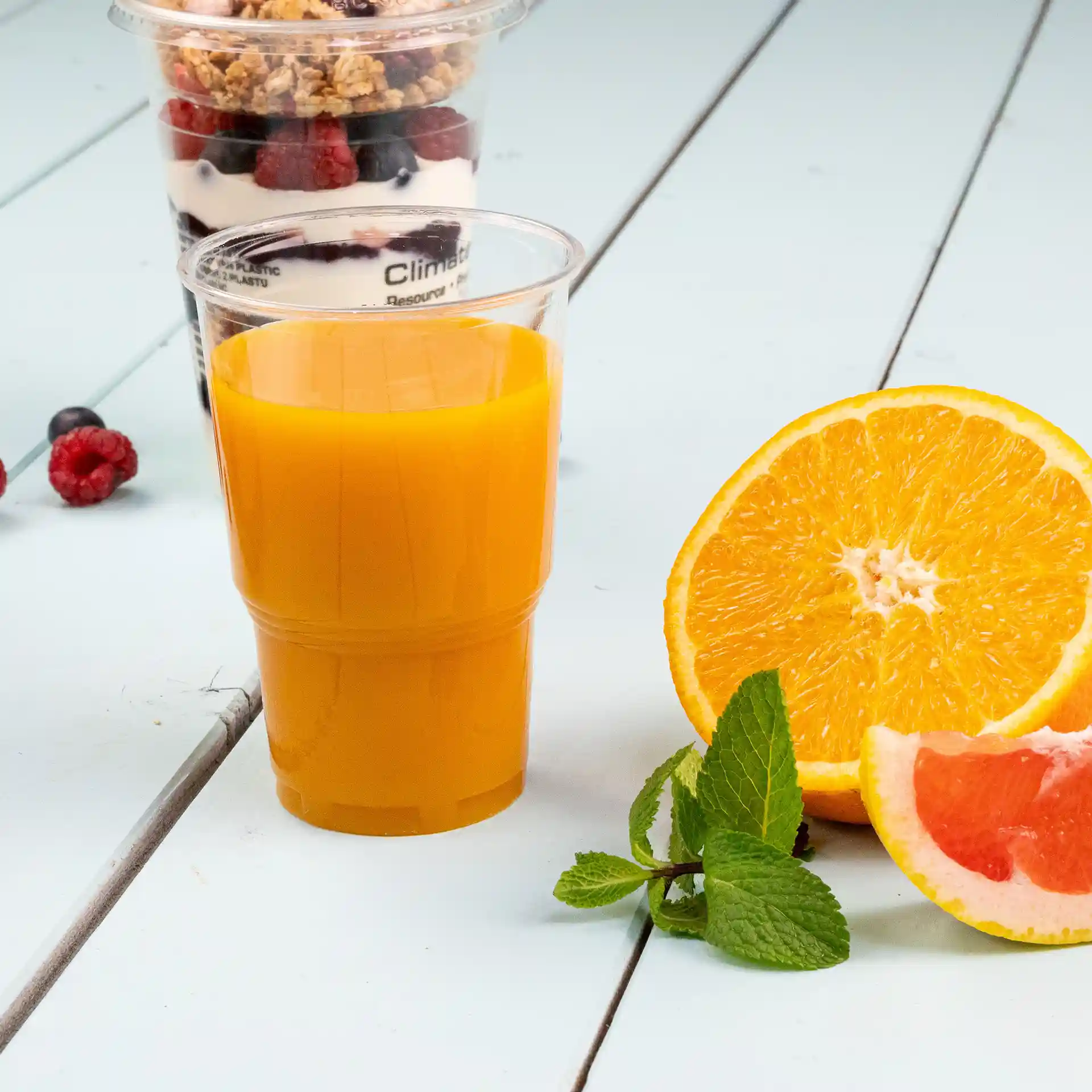 Transparenter Plastikbecher mit Orangensaft, neben einer halbierten Orange und Minzblättern auf einer hellblauen Oberfläche.