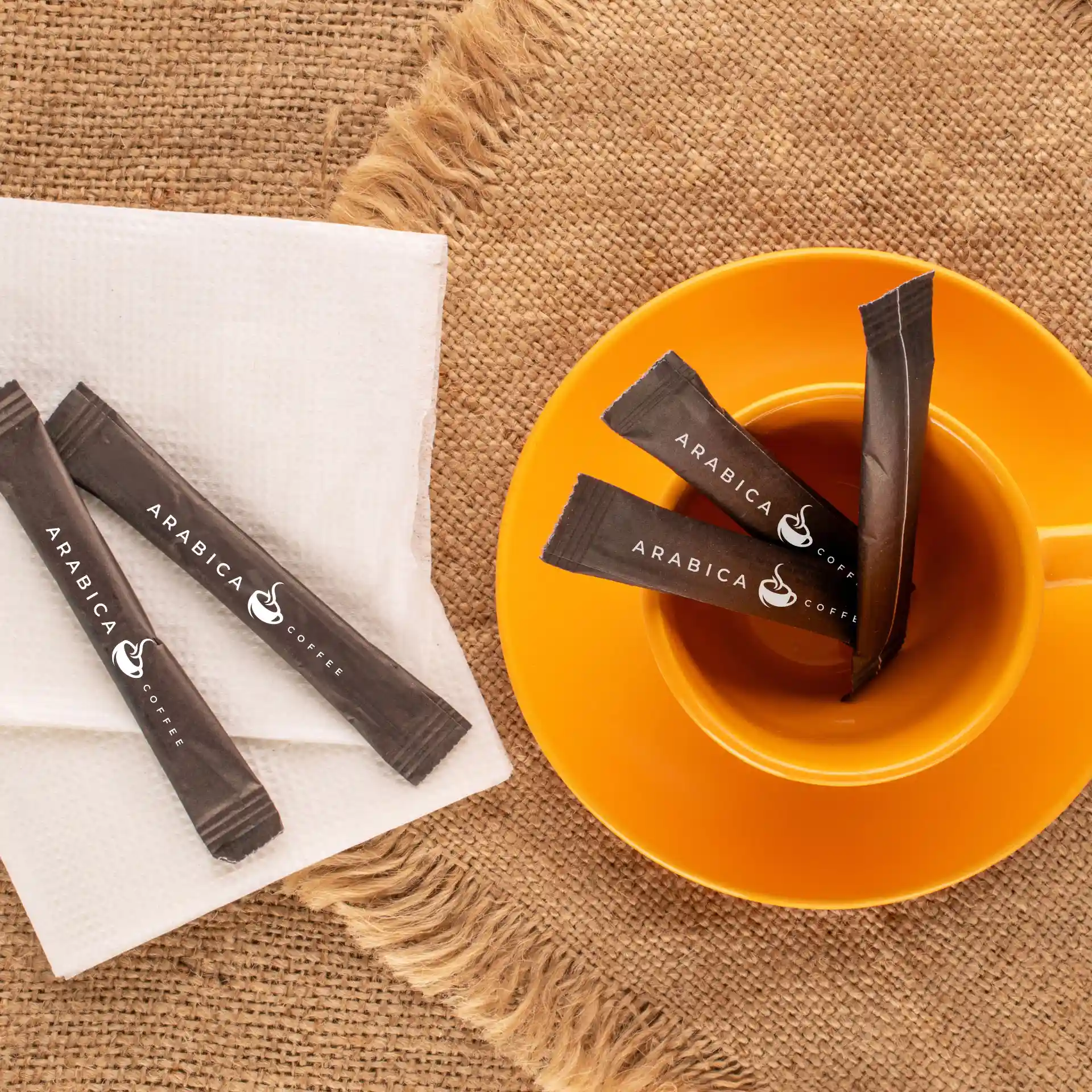 Bild von braunen Zuckersticks mit Logo auf einer weißen Serviette neben einem orangefarbenen Becher auf einer strukturierten Oberfläche.