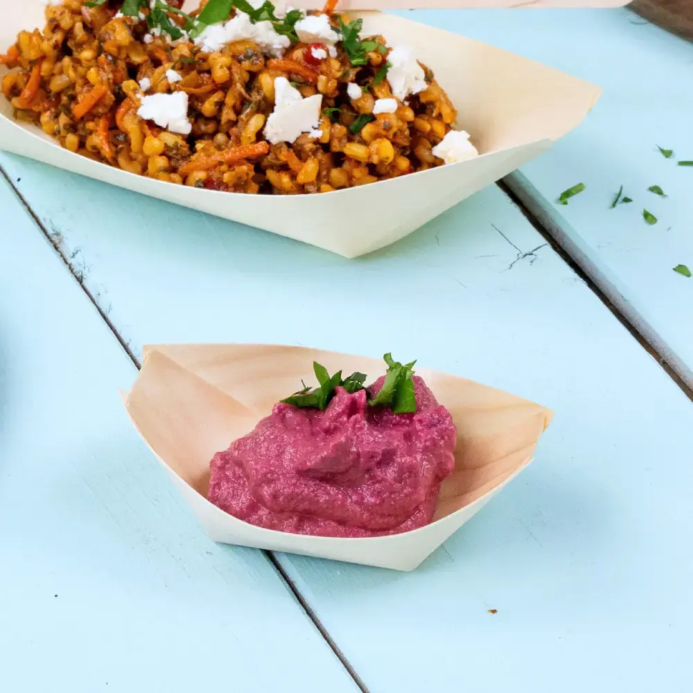 Wooden boat-shaped dish containing pink dip garnished with green herbs, next to a larger dish of mixed grains and vegetables.