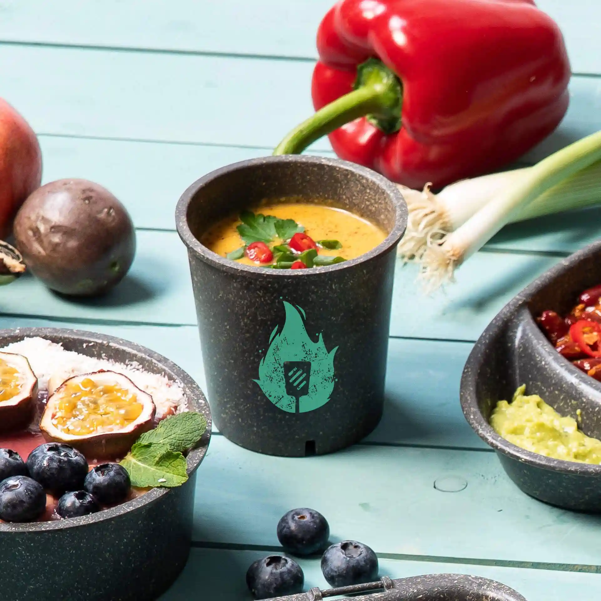 Round grey reusable soup cup with a soup filled to the top, garnished with herbs and chili, on a blue surface with food items.