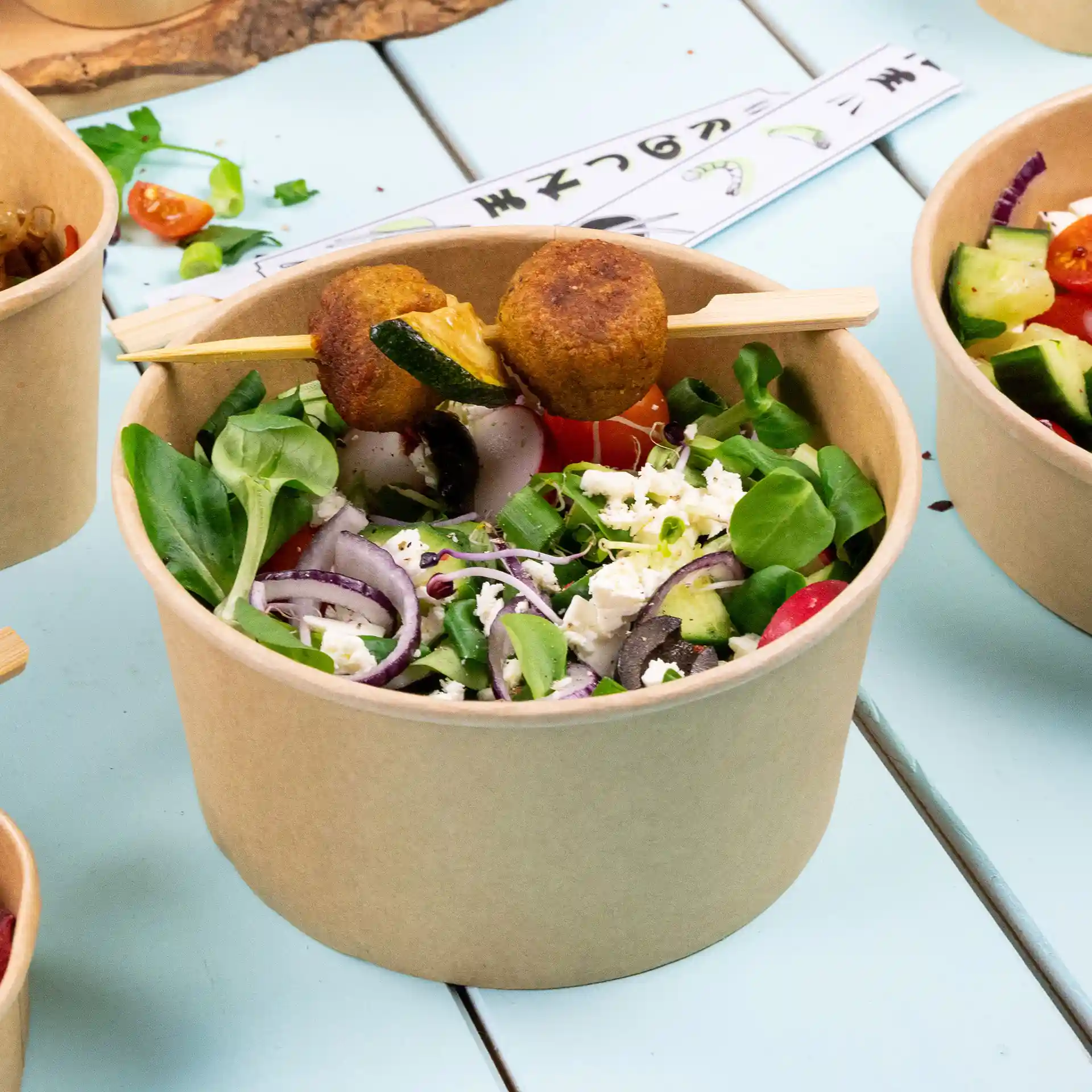 Bamboo bowl filled with mixed salad, topped with two skewered items, on a light blue surface.