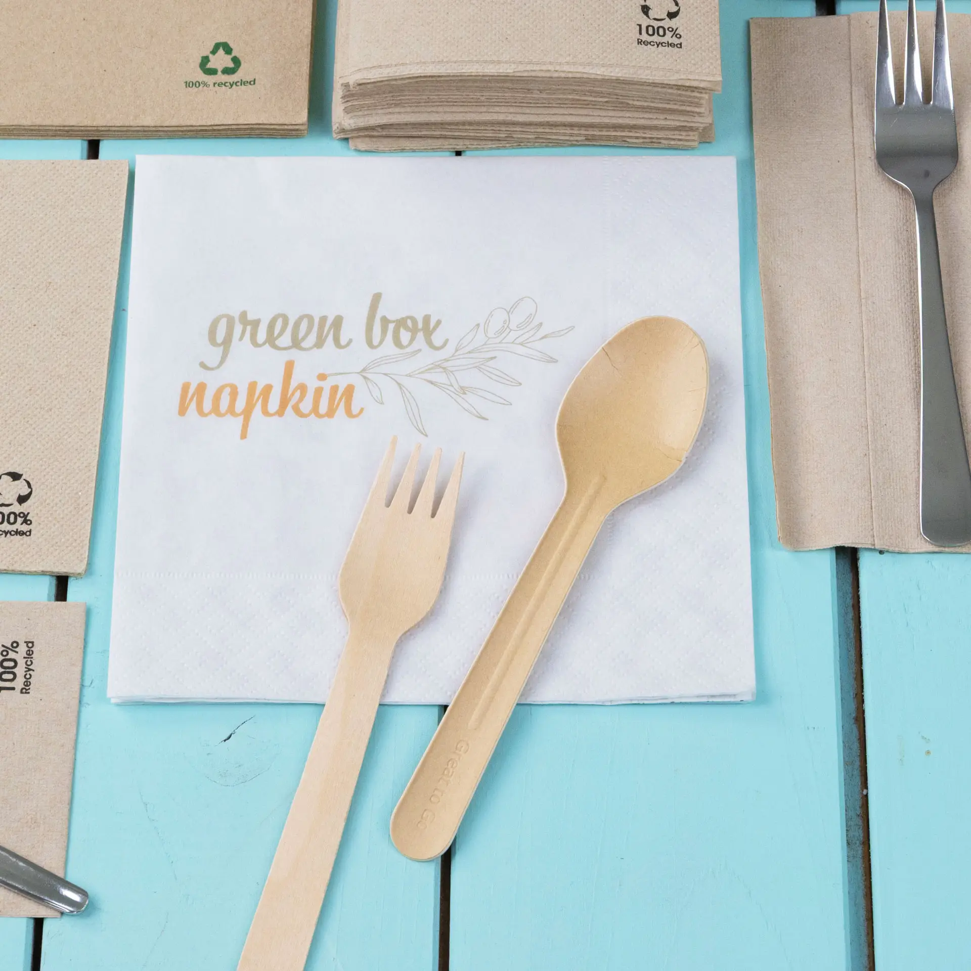 Wooden fork and spoon on a printed lunch napkin labeled "green box napkin," with stacked recycled napkins in the background.