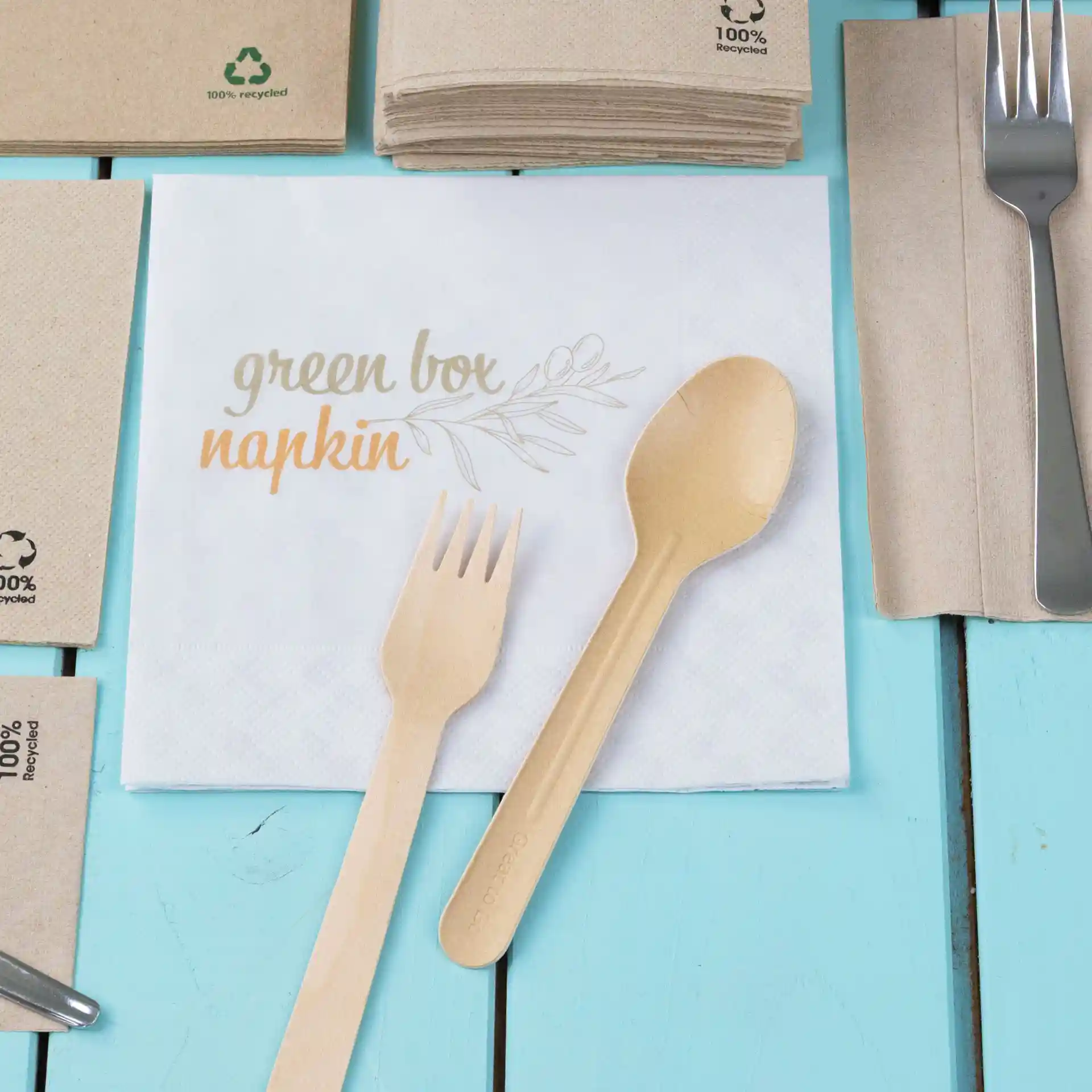 White printed napkin with the text "green box napkin," accompanied by a wooden fork and spoon on a blue surface.