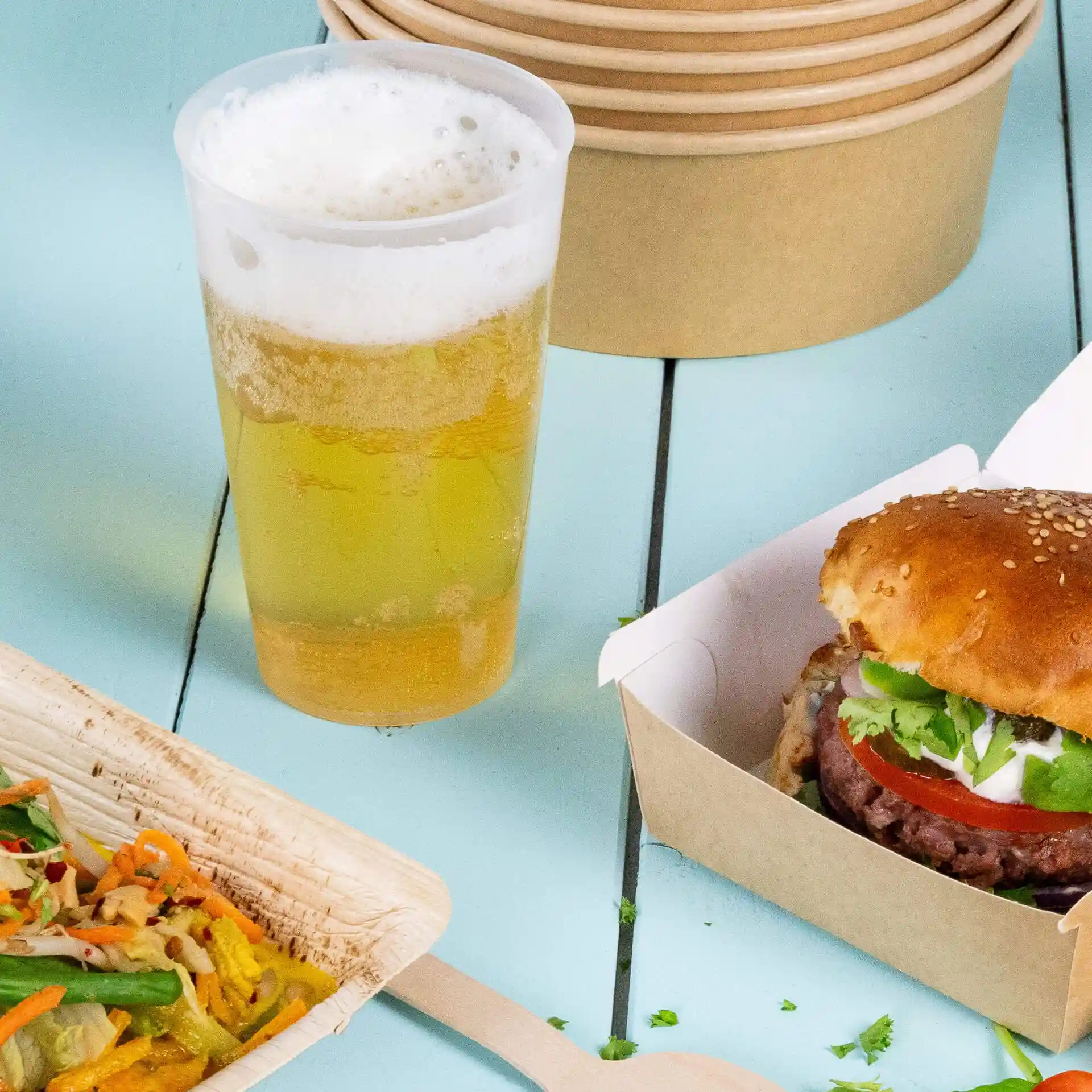 Image of a transparent reusable cup with beer, a sesame seed burger in a box, and a wooden plate with salad.