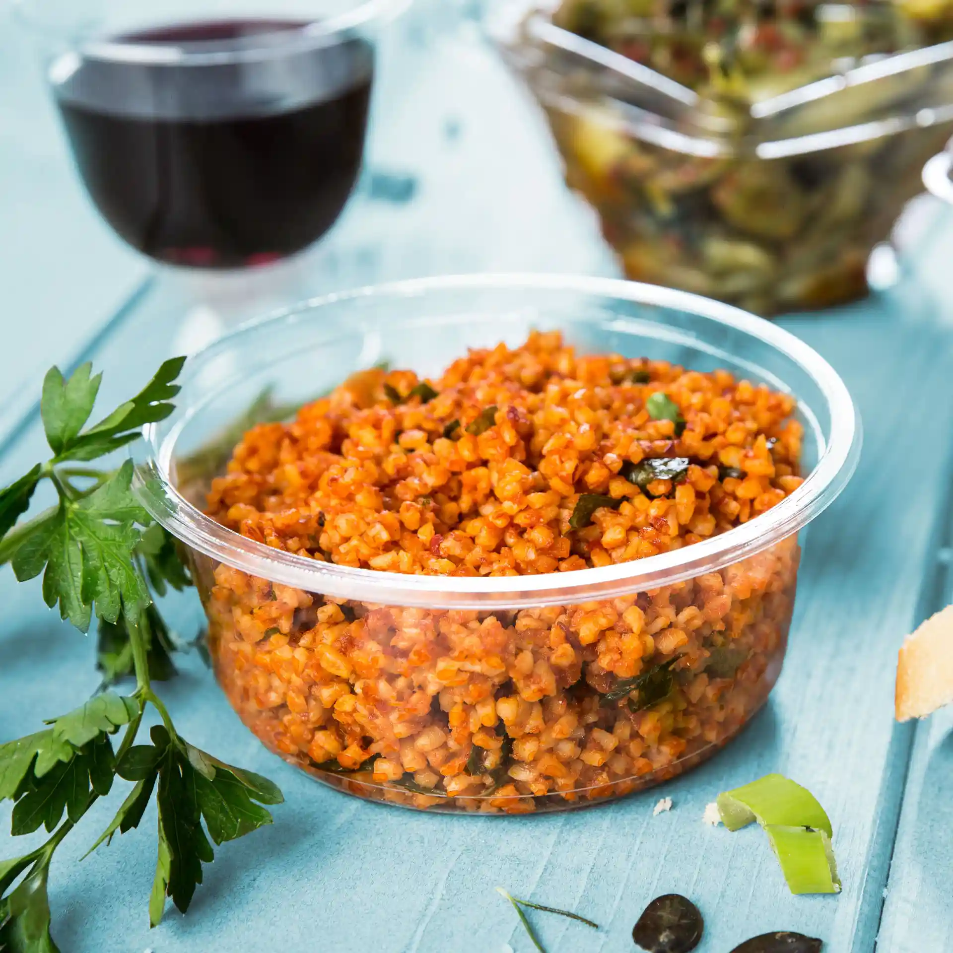 Round transparent deli container with bulgur wheat dish, garnished with herbs, on a blue surface.