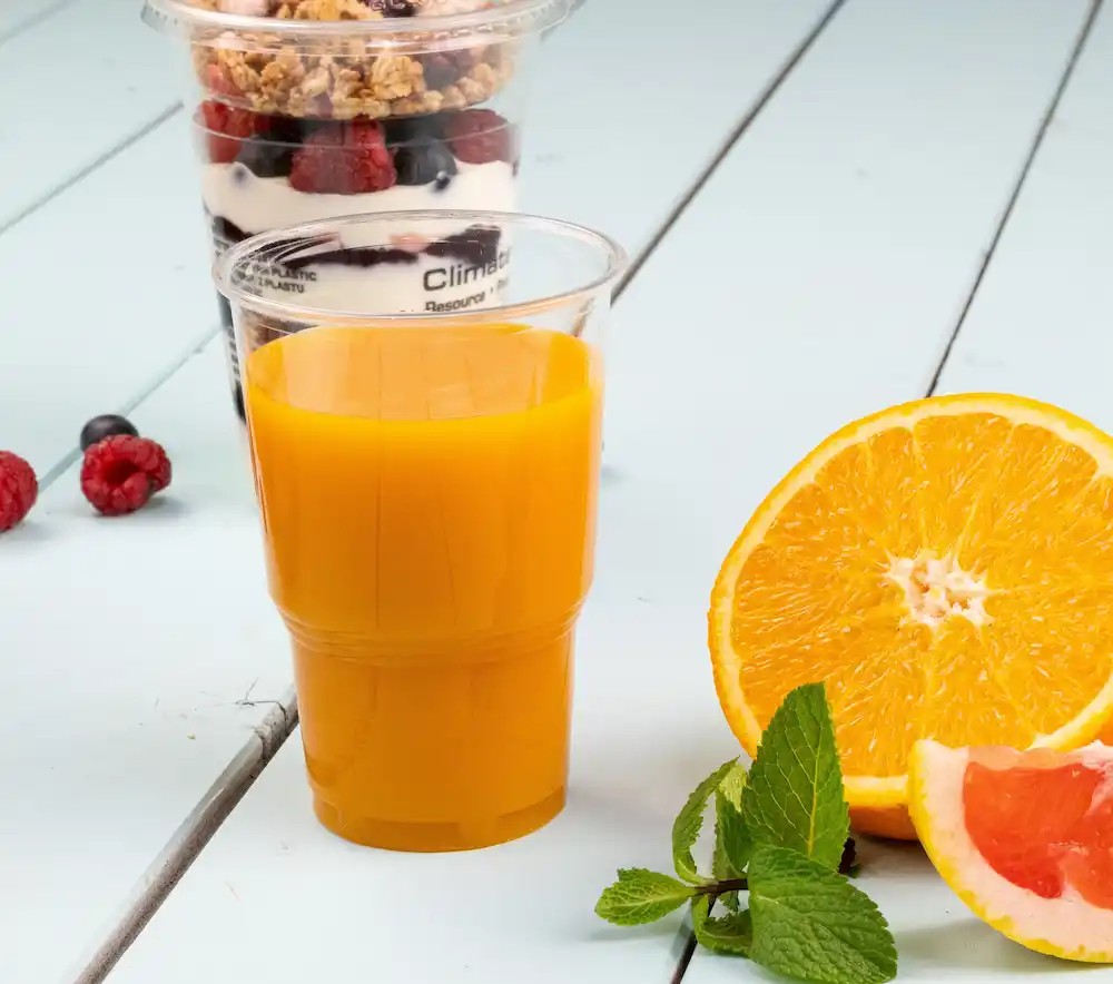 Orange beverage in a transparent cup next to a halved orange and mint leaves, with a dessert cup in the background.