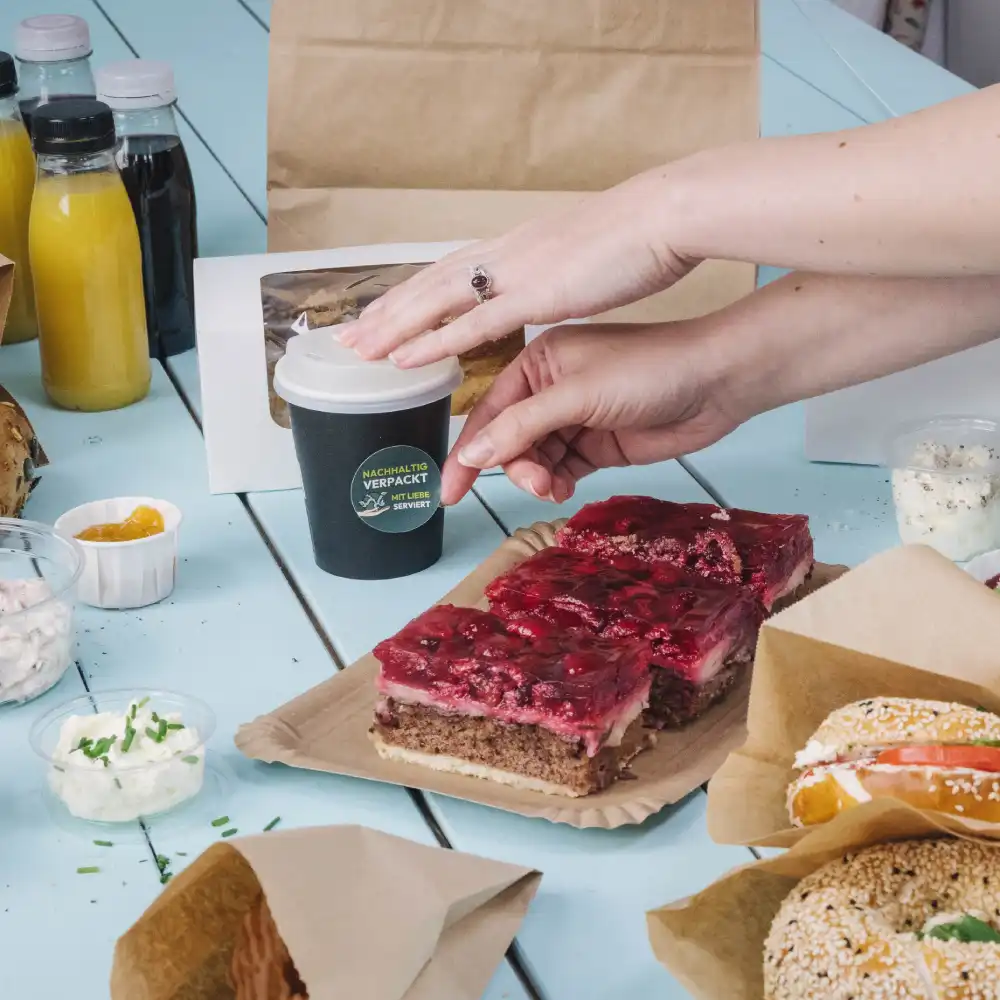 Eine Hand greift nach einem kleinen schwarzen Becher auf einem Tisch mit verschiedenen Lebensmittel; Schichtkuchen, Saucen und Getränke in der Nähe.