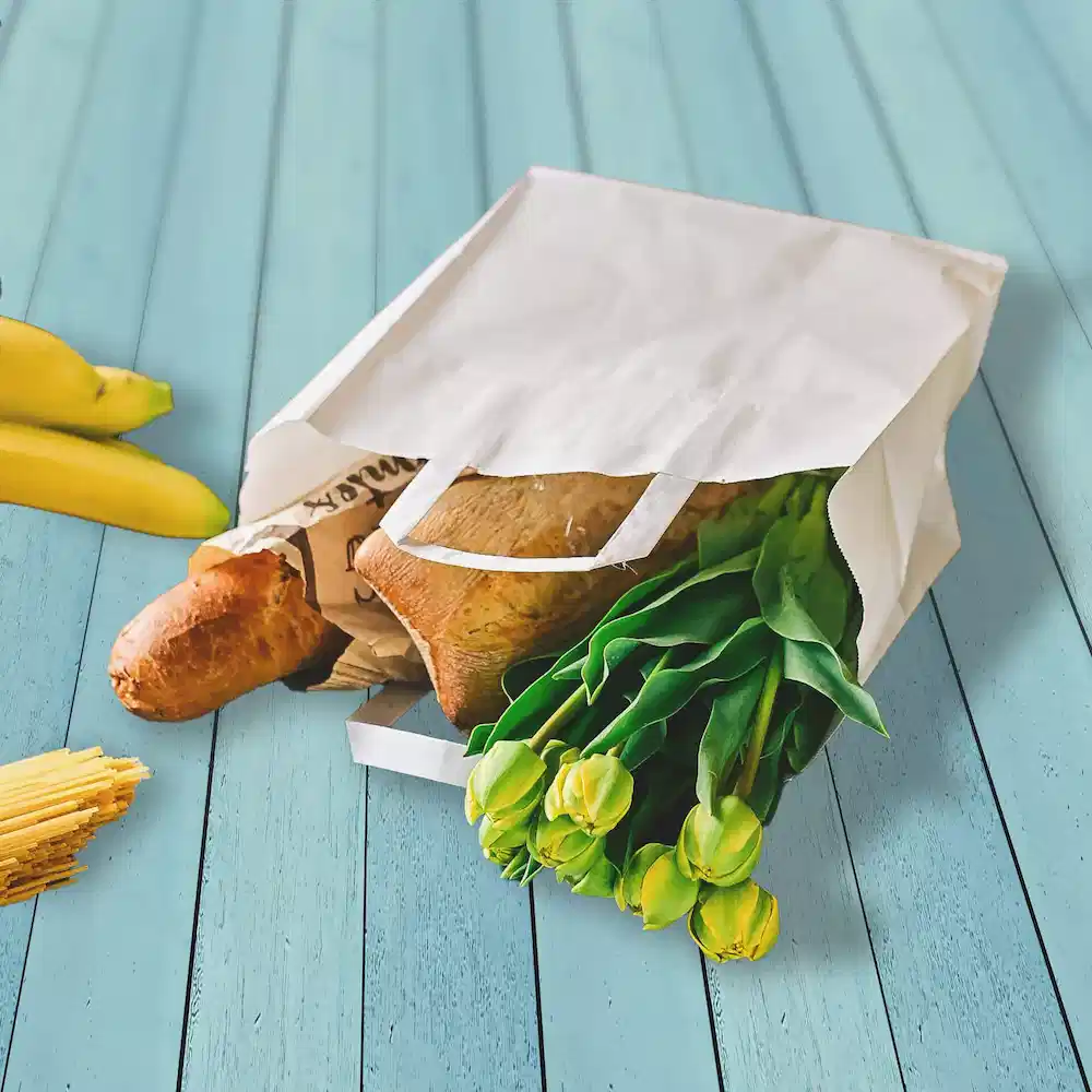 Weißer Papiersack voller Lebensmittel, darunter Brot, Tulpen und Bananen auf einer blauen Holzoberfläche.