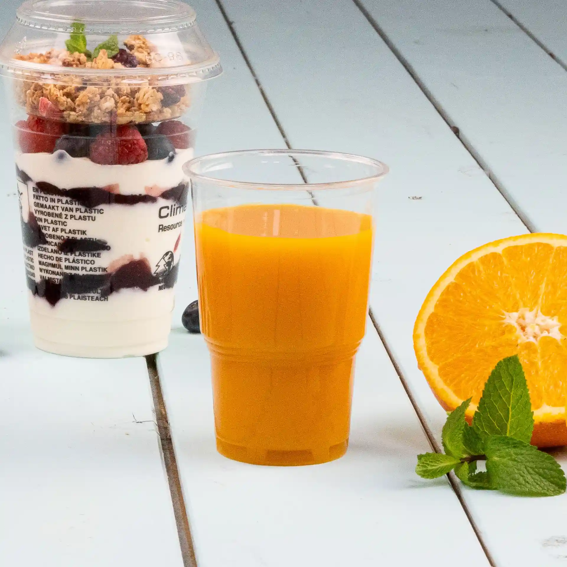 Image of a transparent plastic cup filled with orange juice, placed next to a yogurt parfait and a half orange slice.