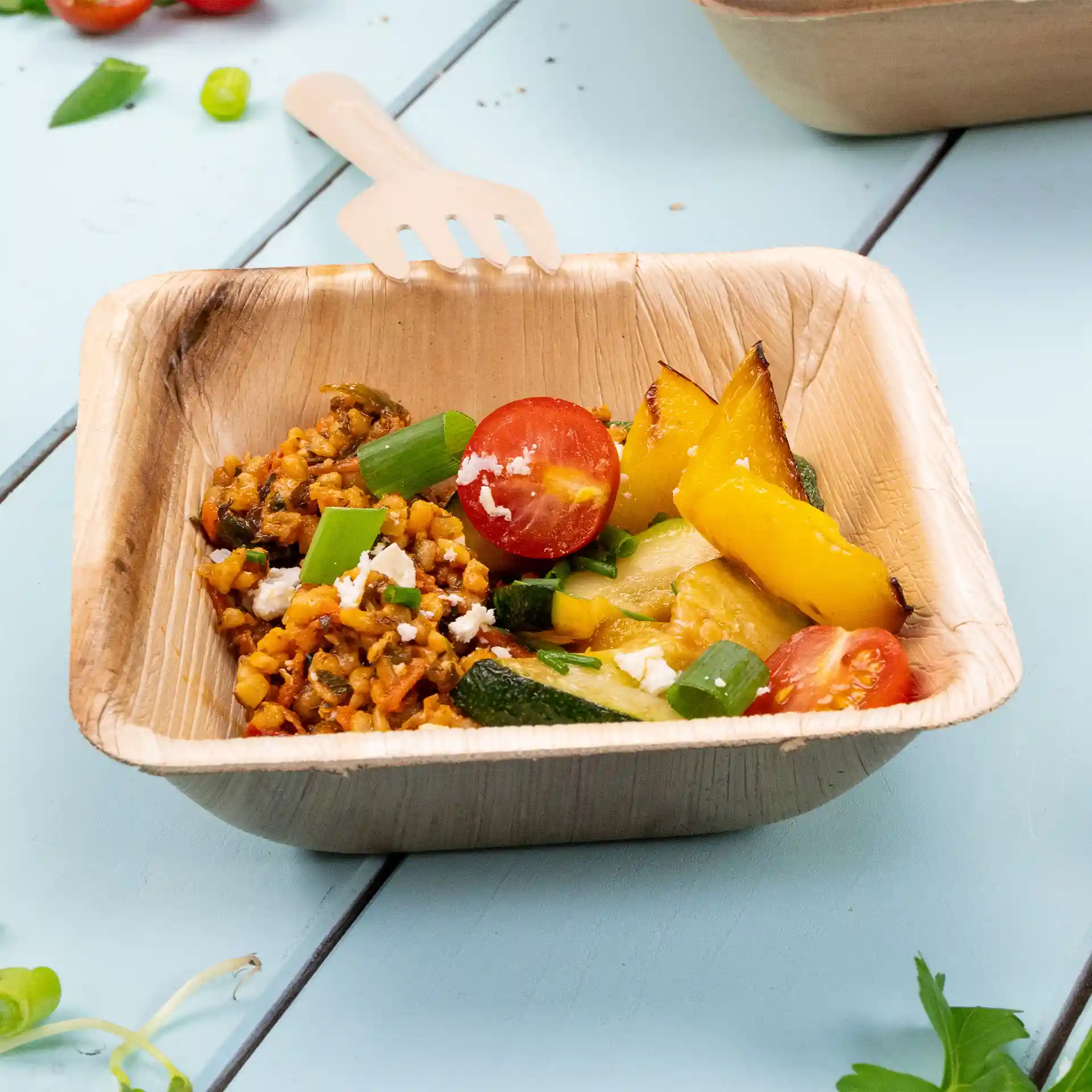 Square palm leaf bowl filled with rice, vegetables, and a cherry tomato, with a wooden fork resting on the edge.