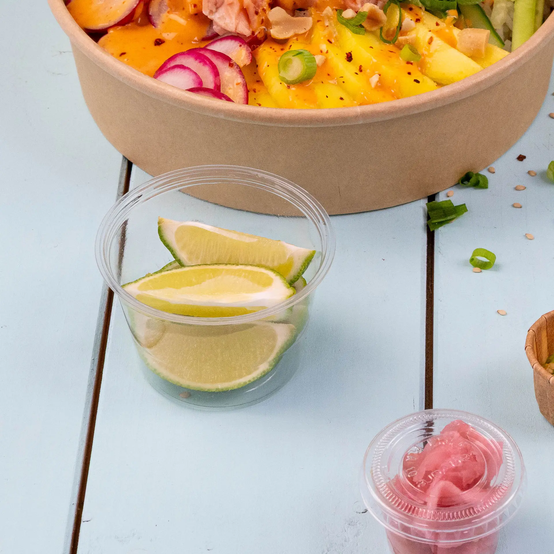 Klare PLA-Becher mit Limettenstückchen und pink eingelegten Zwiebeln, neben einer Schüssel mit Salatzutaten.