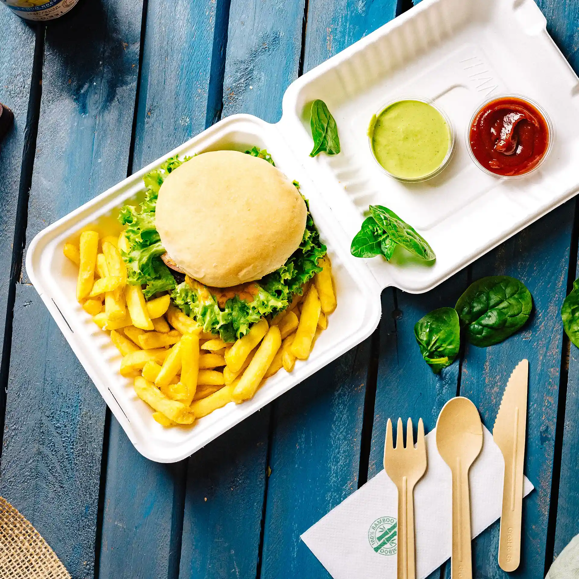 Weißer rechteckiger Behälter mit Klappdeckel, Burger, Pommes, grüner Soße, Ketchup und Holzbesteck auf blauer Oberfläche.