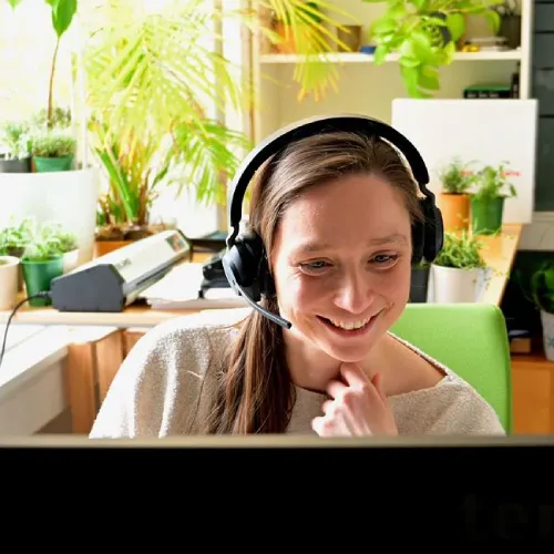 Frau mit Kopfhörern lächelnd am Schreibtisch mit Pflanzen und Computer in einem hellen Raum sitzend.