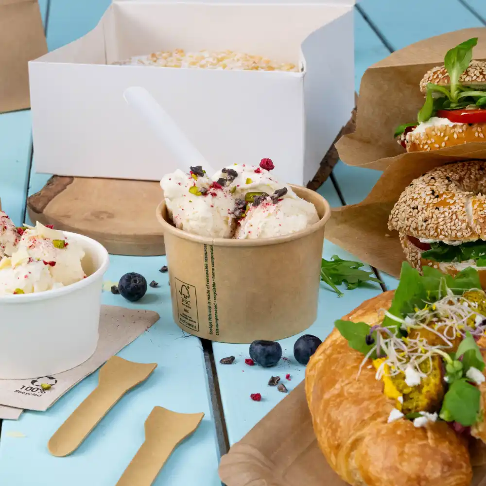 Eiscreme in einem Becher mit Toppings, zwei Sandwiches auf Papier, ein gefülltes Croissant und Blaubeeren auf einer blauen Holzoberfläche.