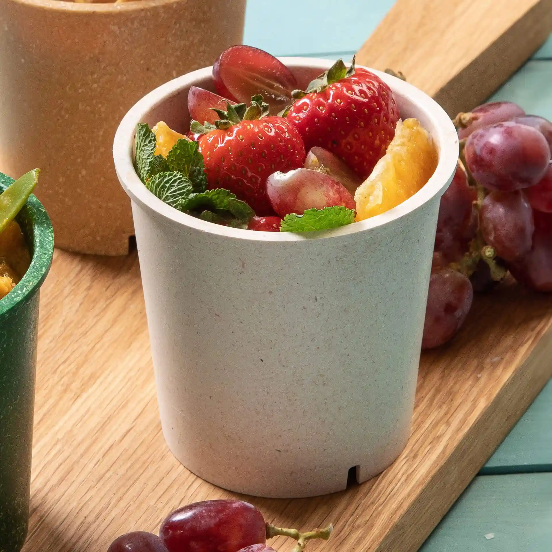 Runder, cremeweißer, wiederverwendbarer Becher, gefüllt mit Erdbeeren, Trauben und Minzblättern auf einer Holzoberfläche.