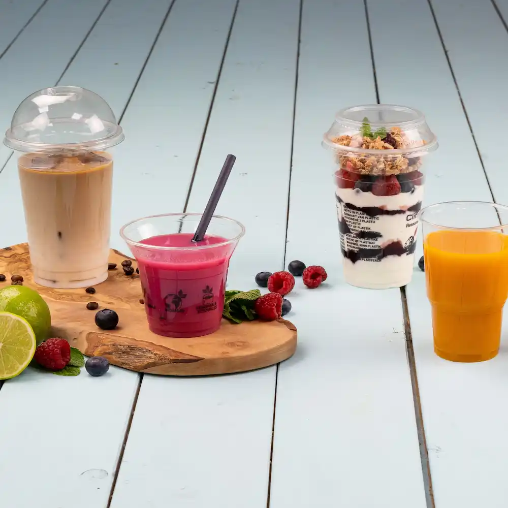 Four drinks on a wooden board: iced coffee in a cup with a dome lid, pink drink with a straw, layered dessert cup, and orange juice.