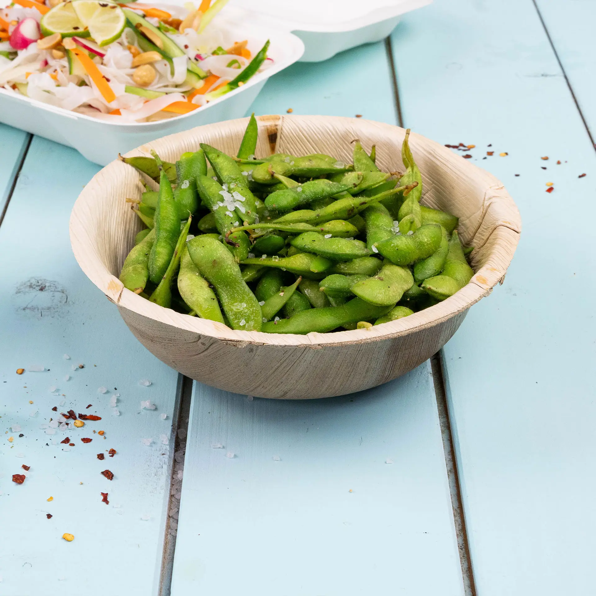 Runde Palmblattschale gefüllt mit grünen Edamame, mit Salz bestreut, auf einer blauen Holzoberfläche.