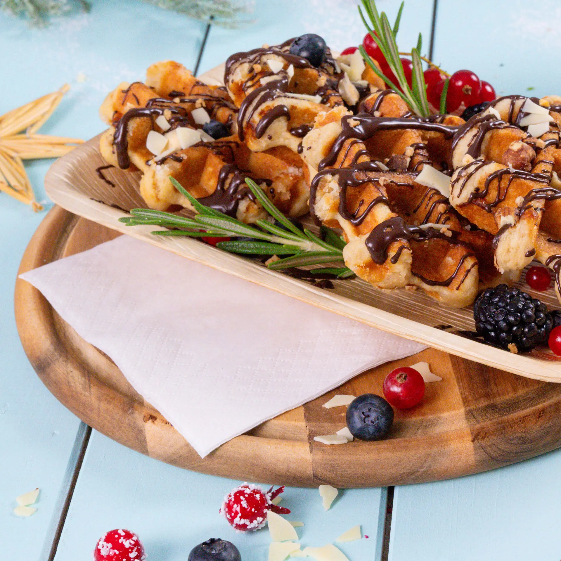 A wooden platter holds waffles drizzled with chocolate, topped with berries and rosemary, beside a white napkin.