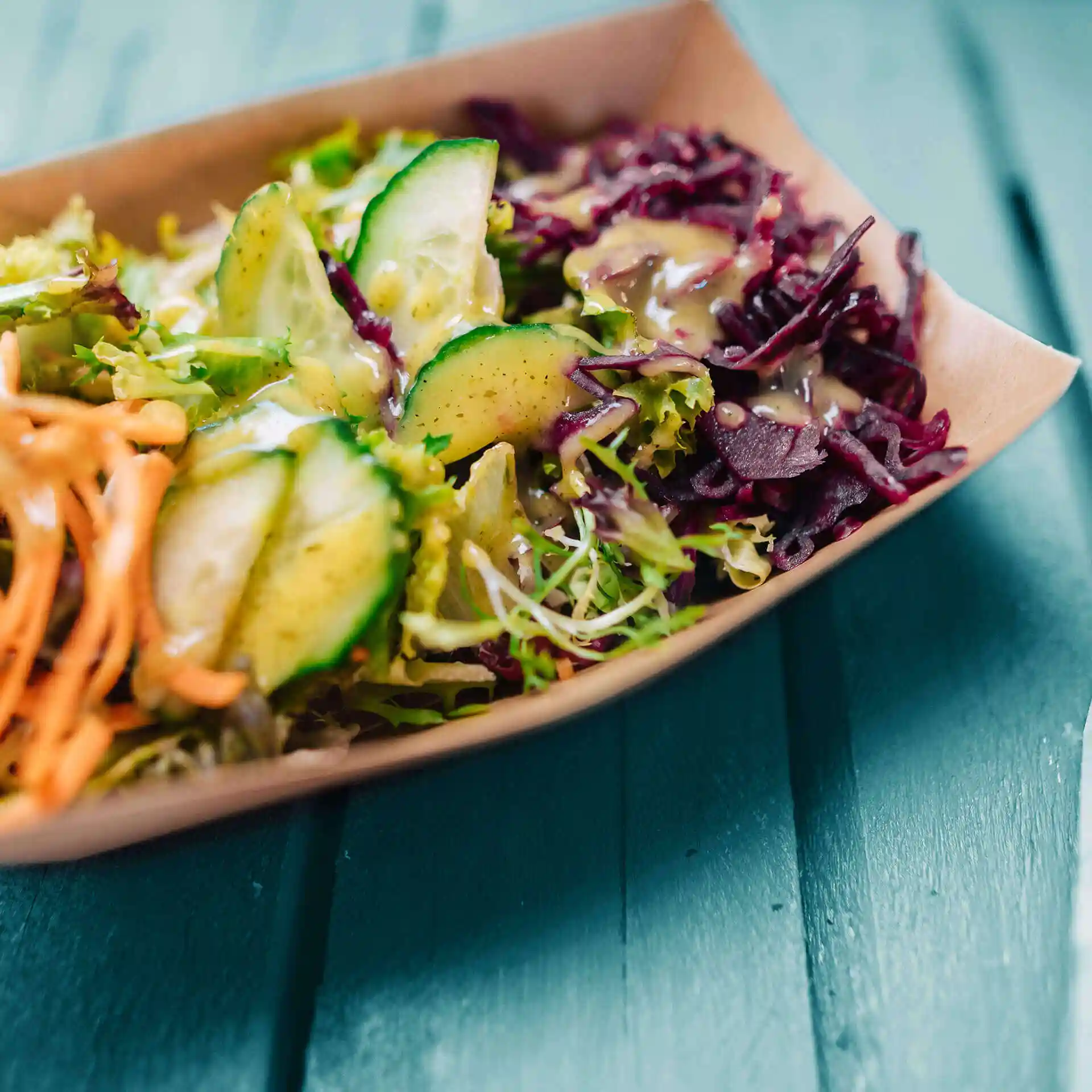 Brown cardboard food tray filled with mixed salad, including cucumber slices and shredded vegetables, topped with dressing.
