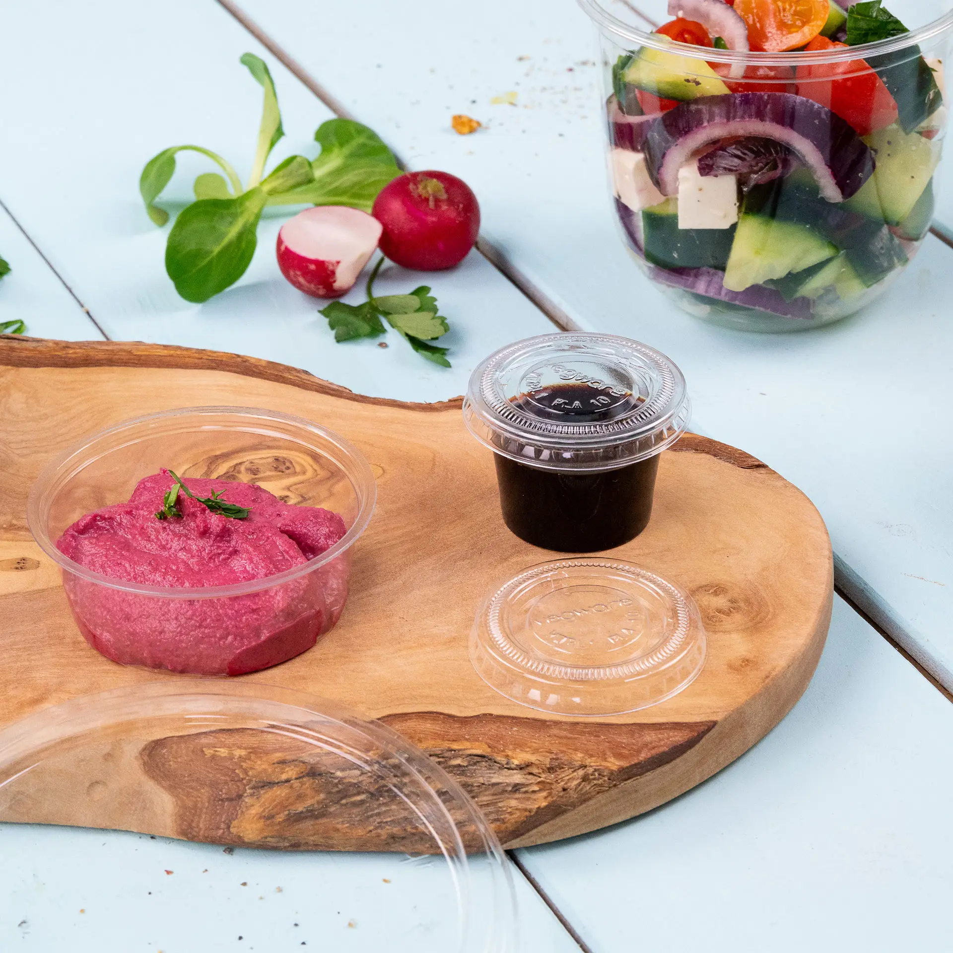 Ein rosa Bioplastik-Portionsbecher gefüllt mit Aufstrich; ein weiterer kleiner Becher mit einer dunklen Sauce; frisches Gemüse im Hintergrund.