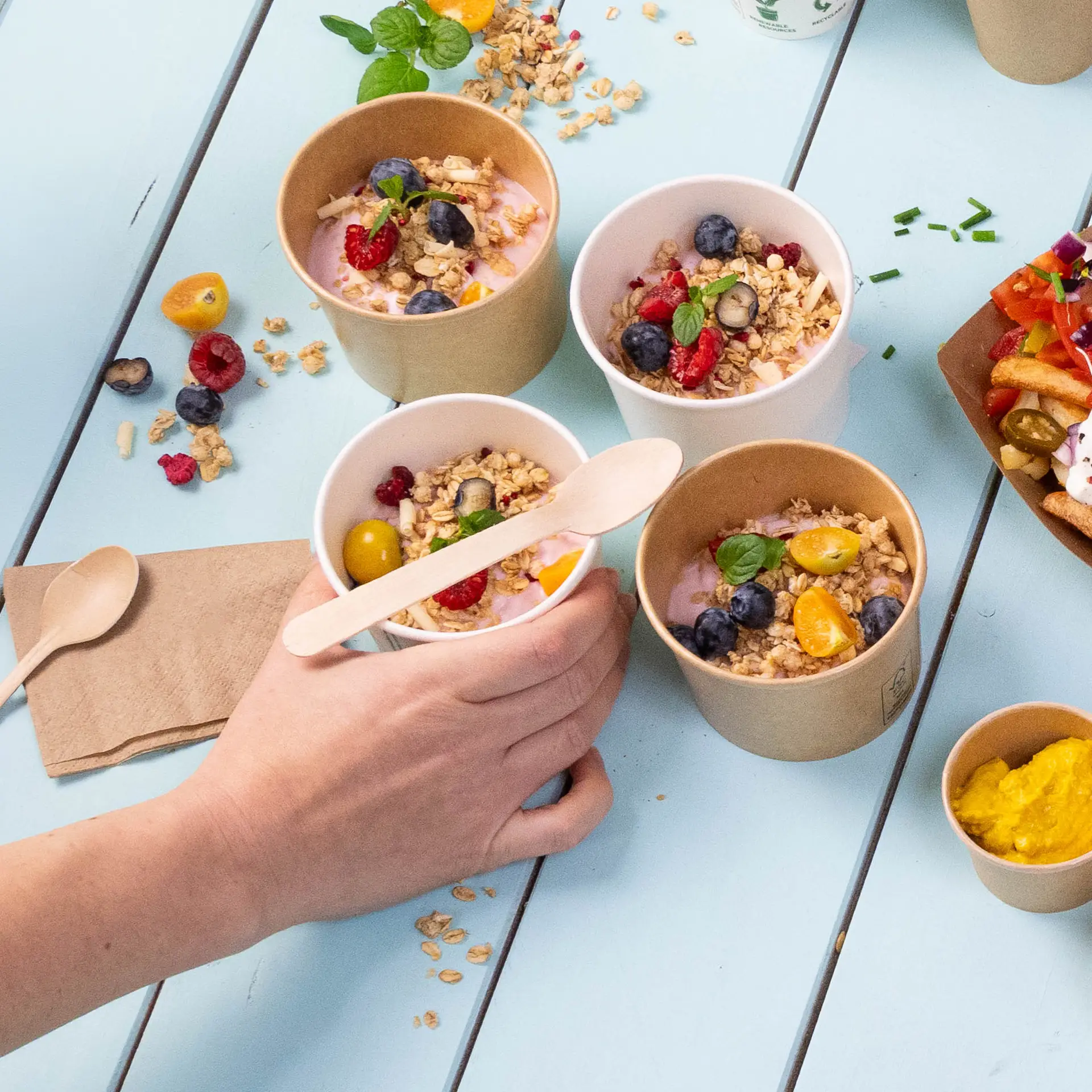 Bild von mehreren kleinen Bechern mit Joghurt, garniert mit Granola und Früchten, eine Hand hält einen Becher, daneben Holzlöffel.