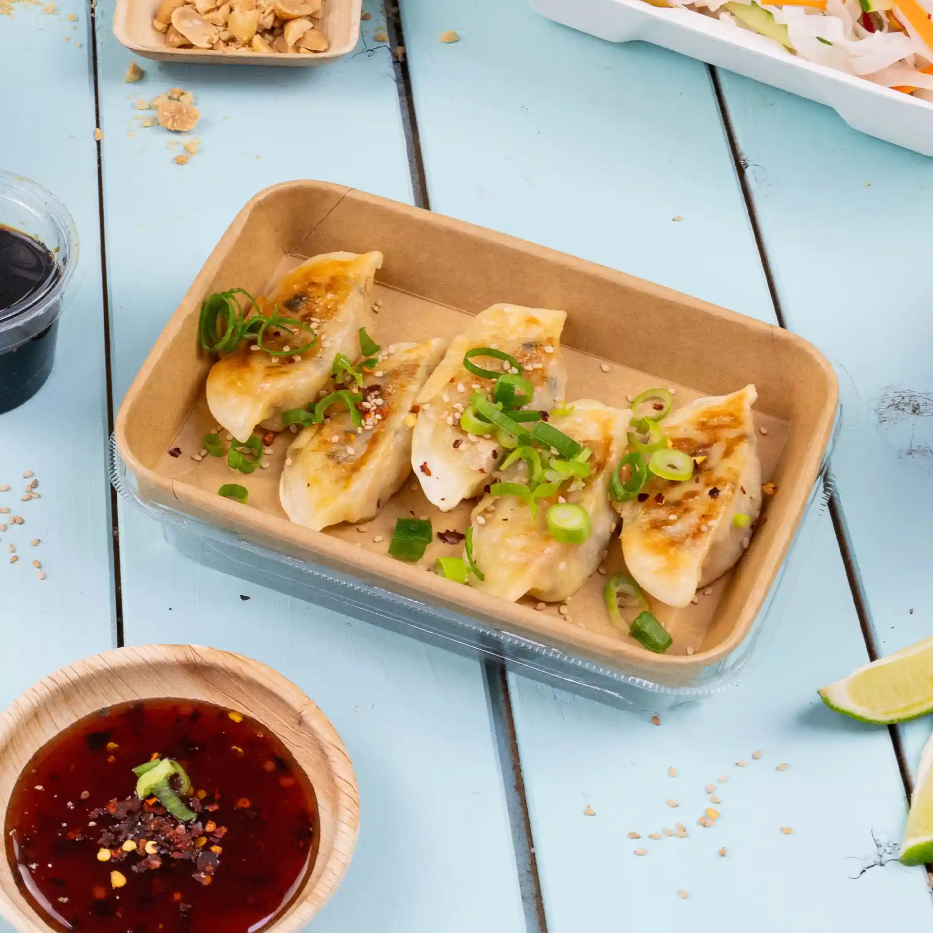 Container with five dumplings topped with green onions and sesame, accompanied by a bowl of dipping sauce.
