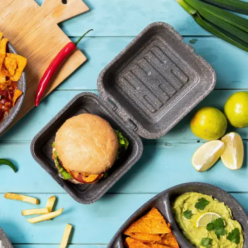 Burger in einer wiederverwendbaren Kraftbox mit Salat und Saucen, umgeben von Chips, Pommes, Guacamole und Limettenstücken.