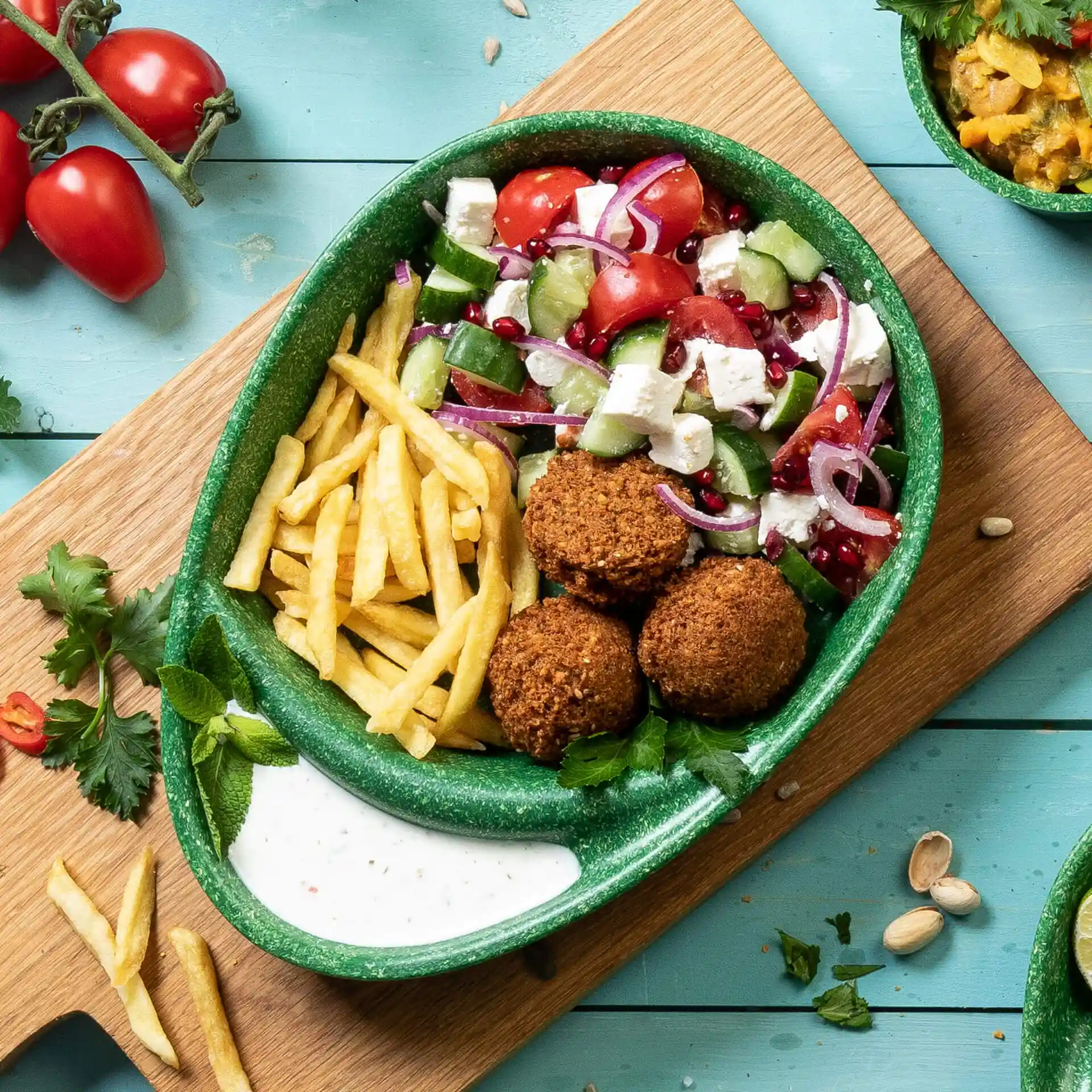 Green divided plate with falafel balls, fries, salad, and sauce, placed on a wooden surface with tomatoes and herbs nearby.