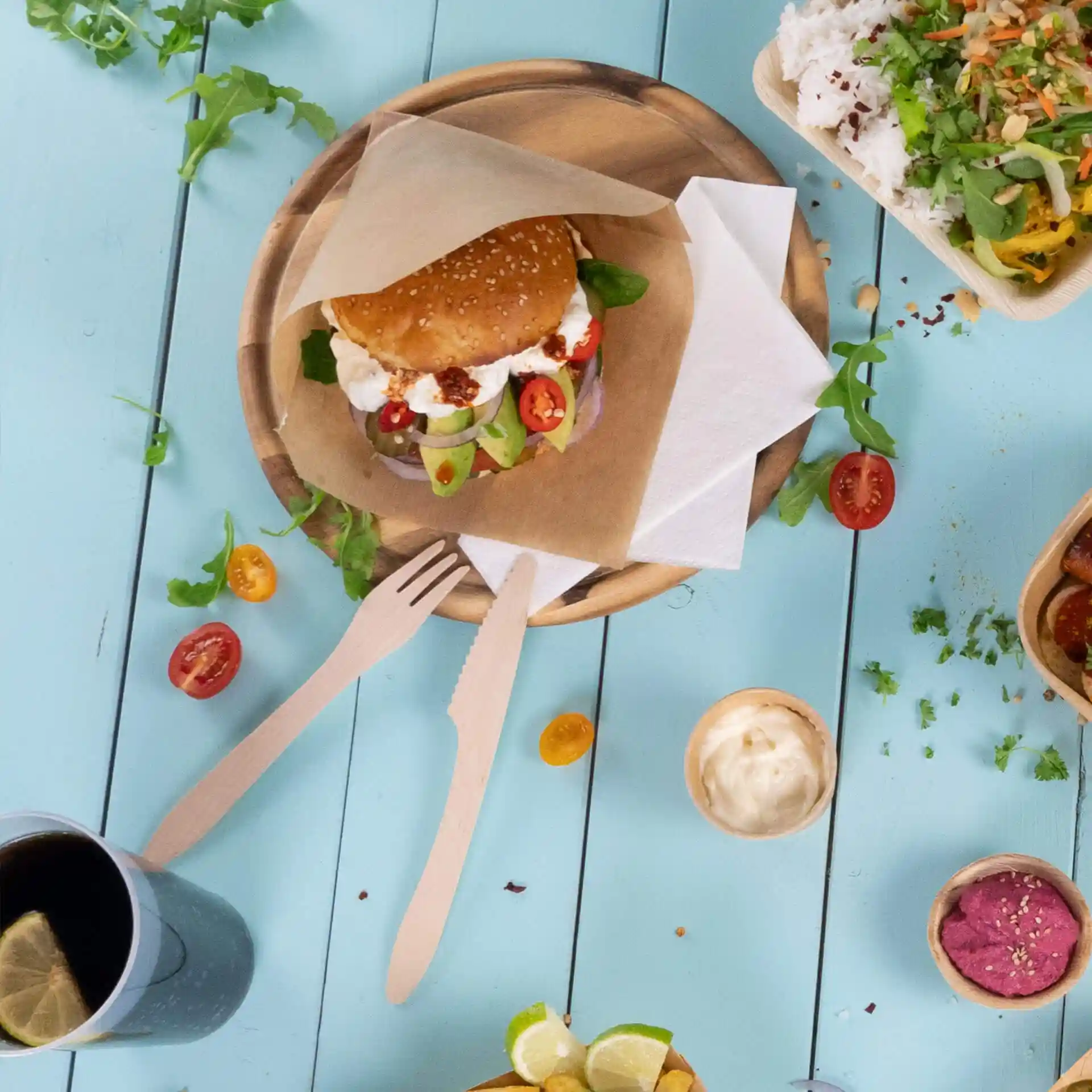 Bild eines Sesambrötchen-Sandwichs mit Gemüse auf einem Holzbrett, begleitet von Holzutensilien und einem Getränk.
