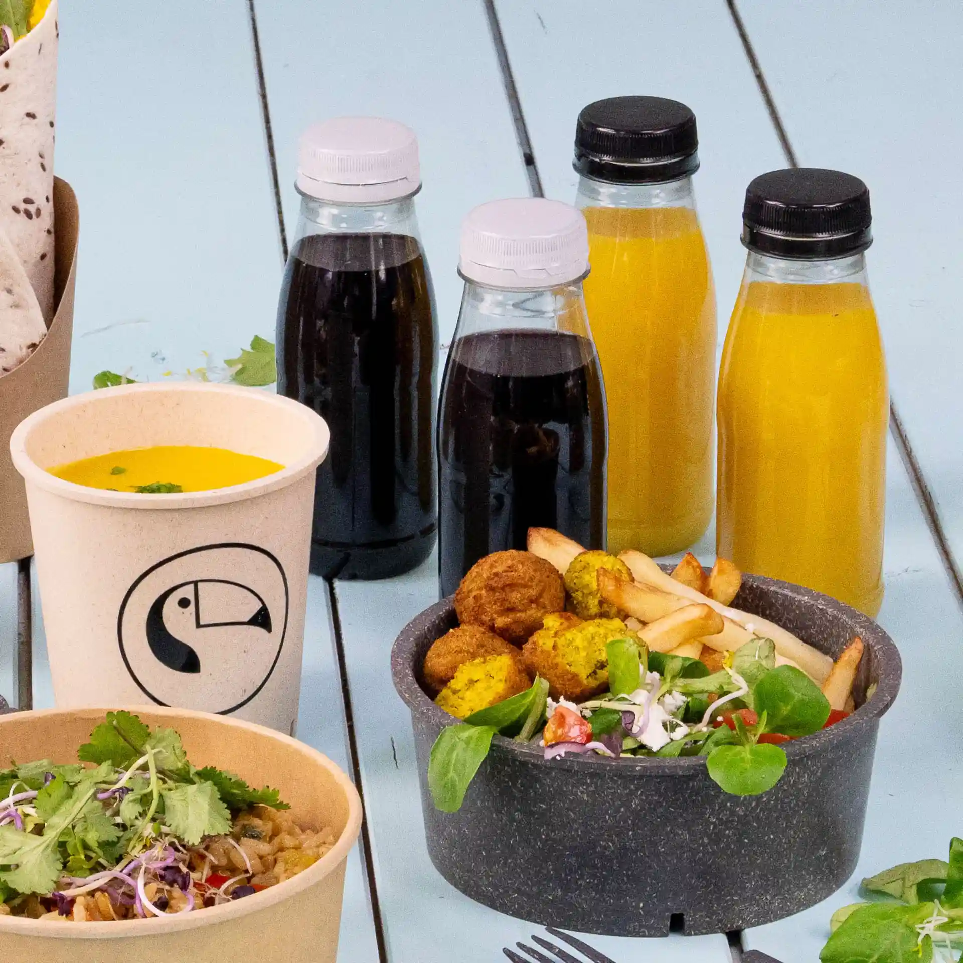 Bild eines Essensarrangements mit einem Becher Suppe, zwei Schalen mit Salat und Falafel sowie vier klaren Flaschen mit schwarz-weißen Deckeln.