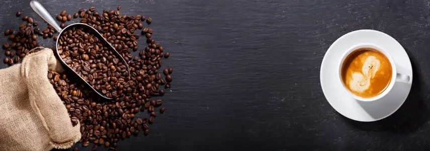 Coffee beans spilled next to a metal scoop and a cup of espresso on a dark surface.