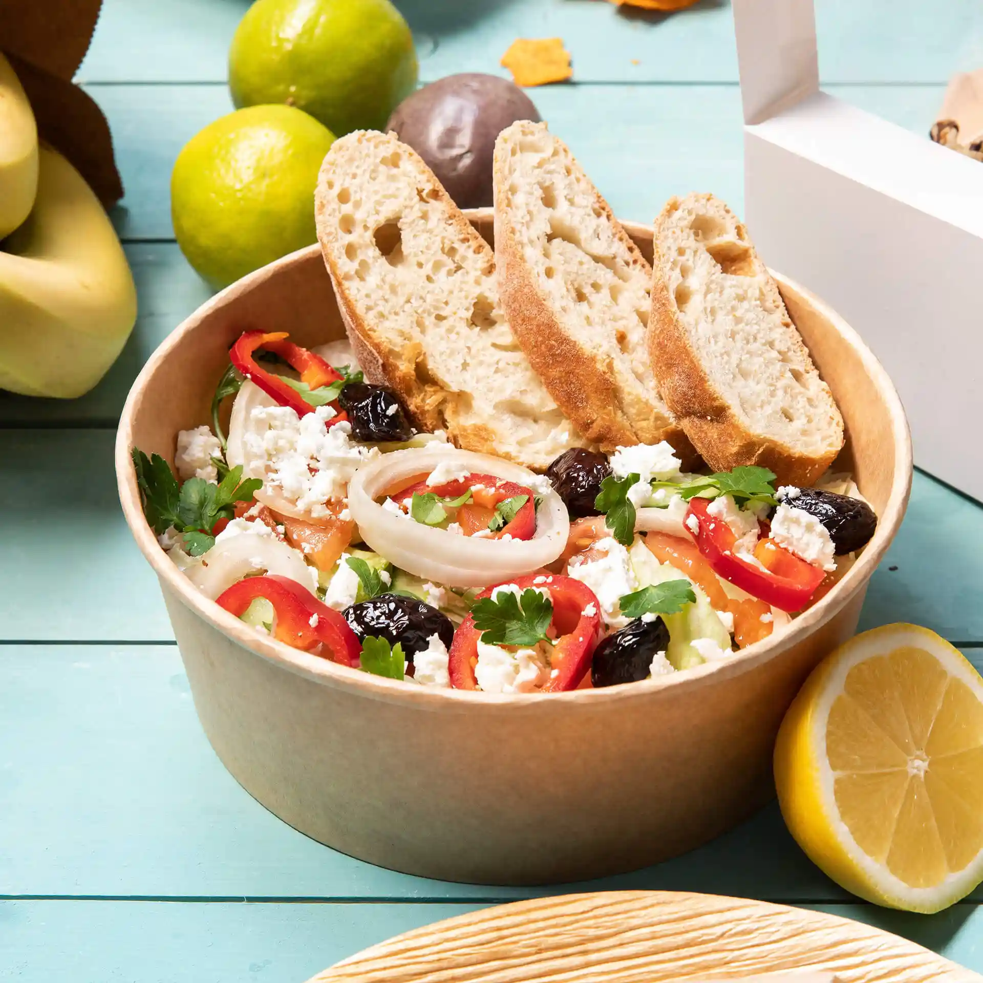 Schüssel mit Salat, belegt mit geschnittenem Gemüse und Feta-Käse, dazu zwei Scheiben Brot, auf einer blauen Oberfläche.