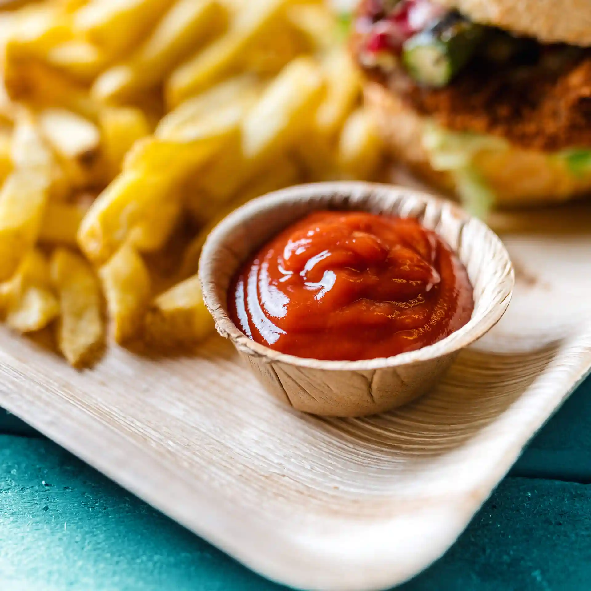 Schüssel aus Palmblatt mit Ketchup, im Hintergrund Pommes auf einer Holzoberfläche.