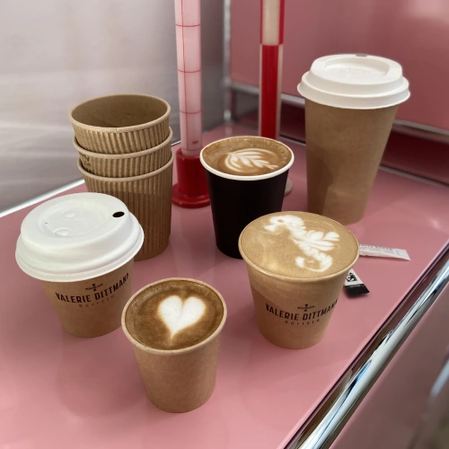 Vielfalt an Bechern, darunter Papier und Plastik, mit Latte-Art-Designs, auf einer rosa Oberfläche angeordnet.