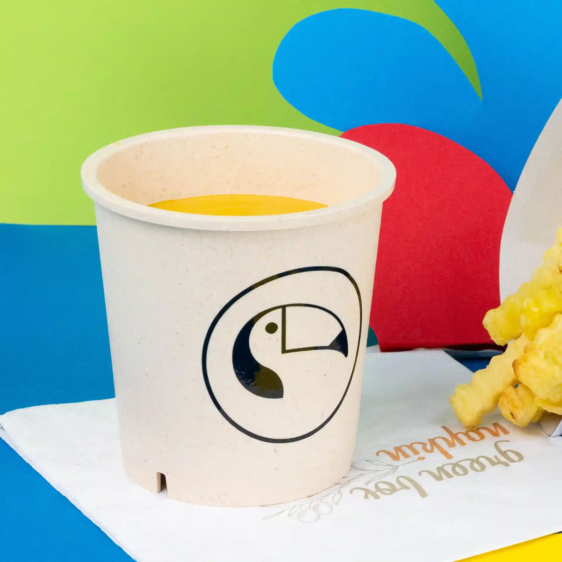 Runde cremeweiße wiederverwendbare Suppenschale mit einem Tukan-Logo, gefüllt mit gelber Suppe, auf einer Serviette.