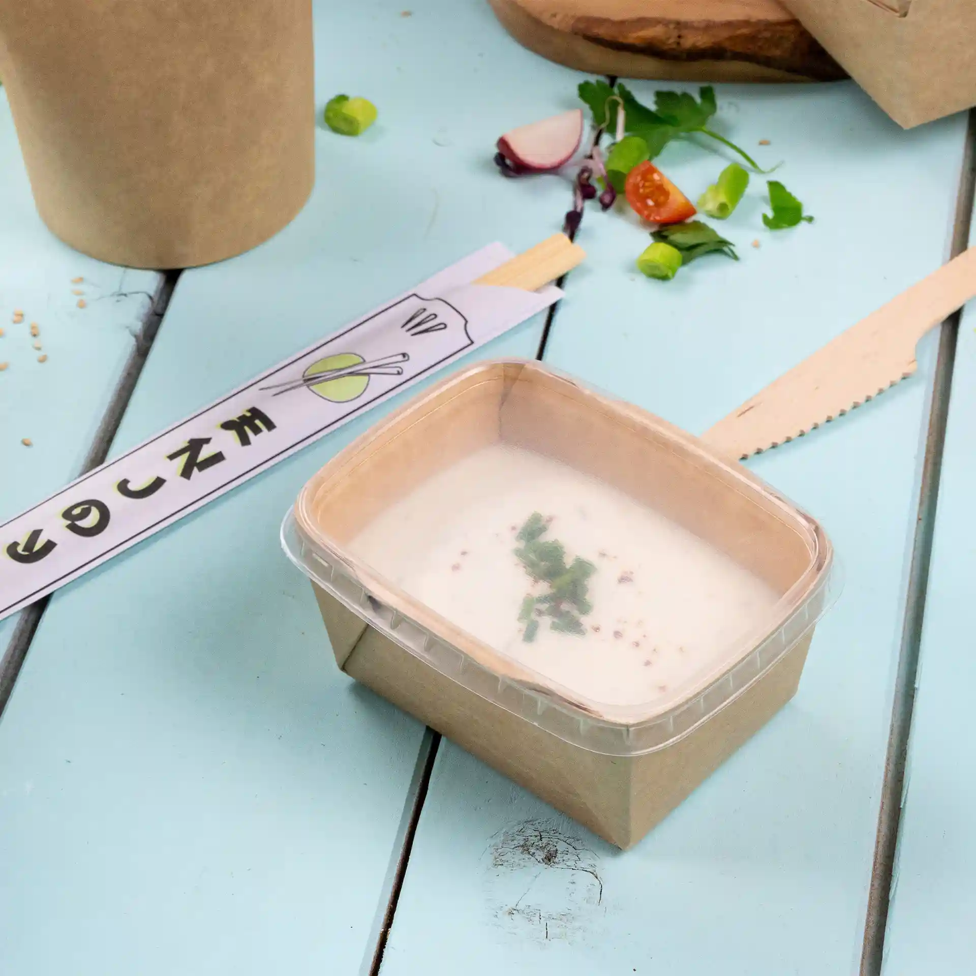 Rectangular clear container with a lid, holding a creamy soup topped with herbs, on a blue wooden surface.