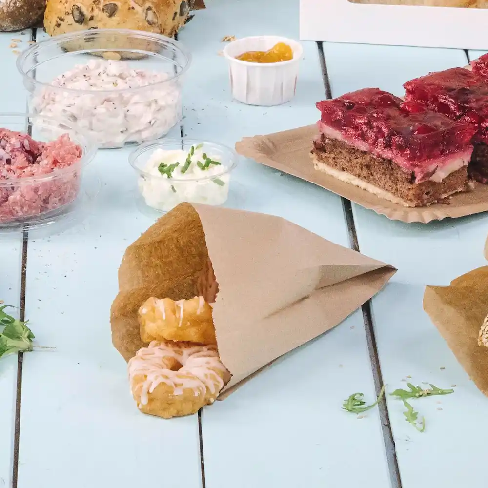 Brown paper bag containing two iced donuts, surrounded by various food items and toppings on a blue tabletop.