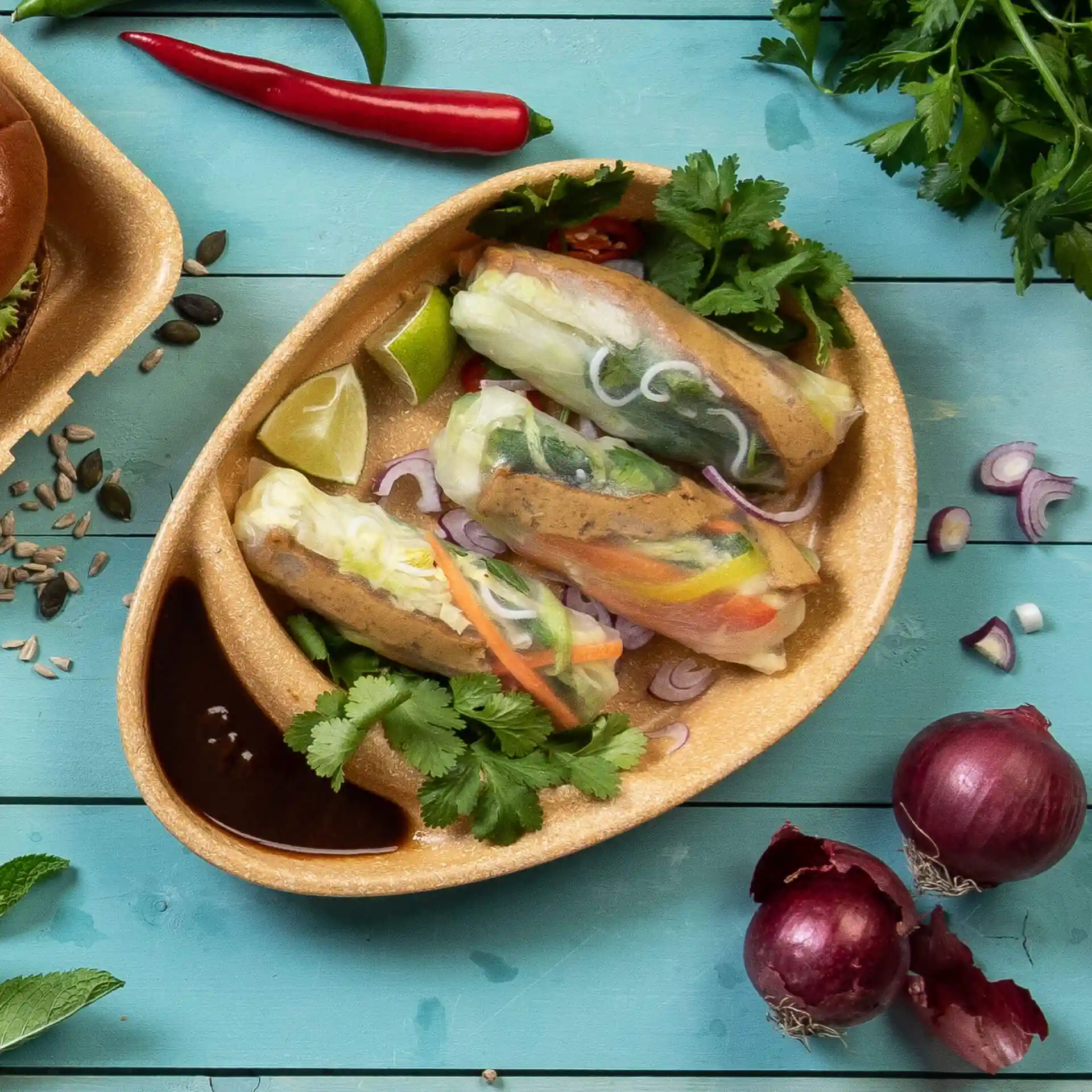 Reusable divided plate with two compartments, containing spring rolls, lime, cilantro, and dipping sauce on a blue background.