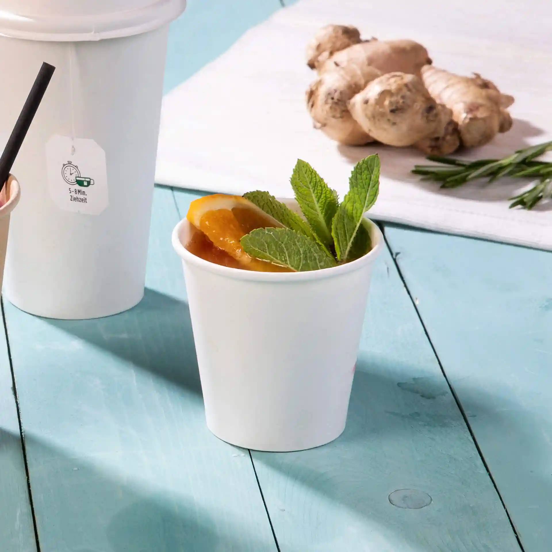 White paper cup filled with a drink, garnished with mint leaves and orange slices, on a blue wooden surface.