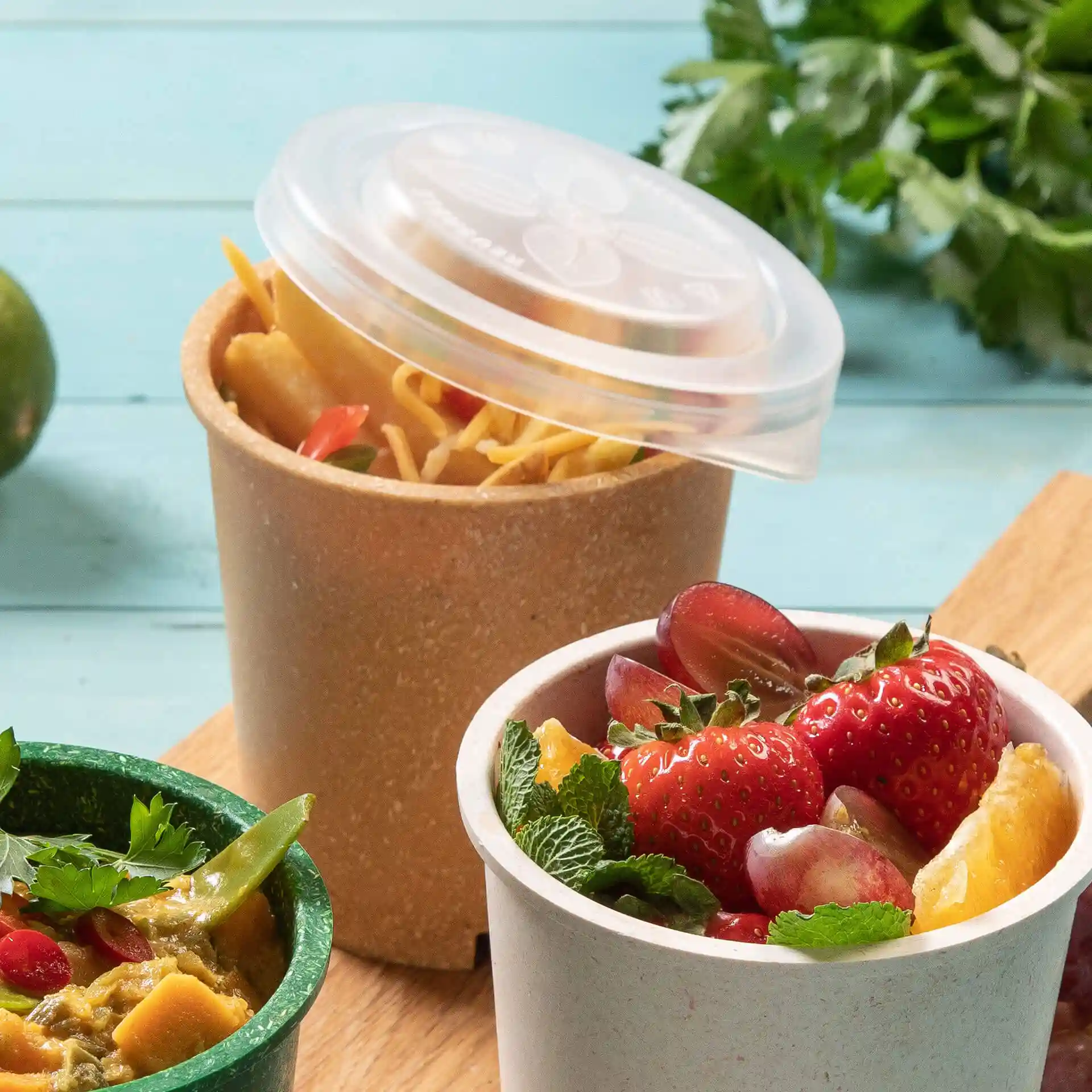 Image of two round cups, one with a transparent lid, containing noodles and the other with fruit, on a wooden surface.