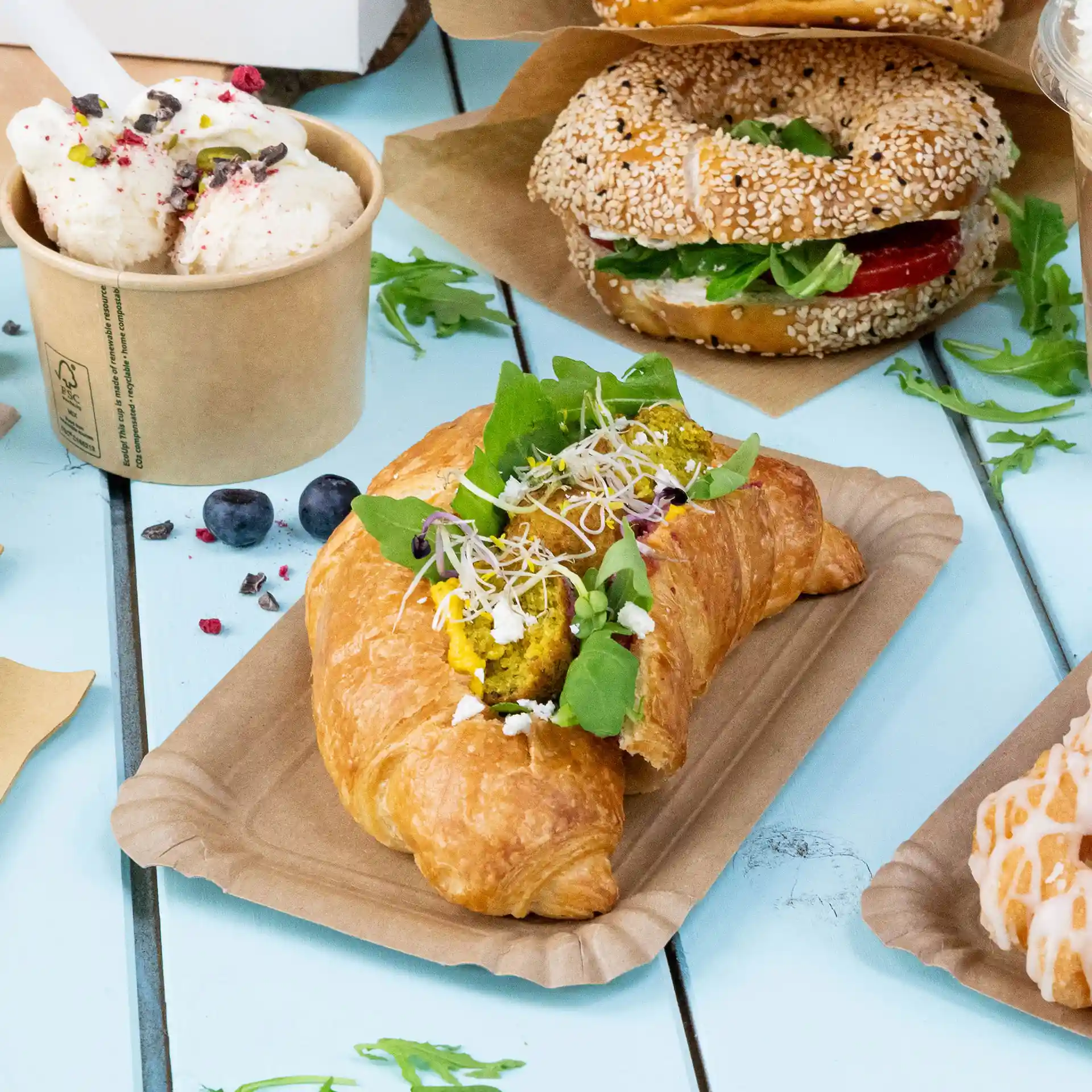 Croissant gefüllt mit grünen Blättern, Sprossen und gelber Füllung auf einem braunen Pappteller, mit anderen Lebensmitteln im Hintergrund.