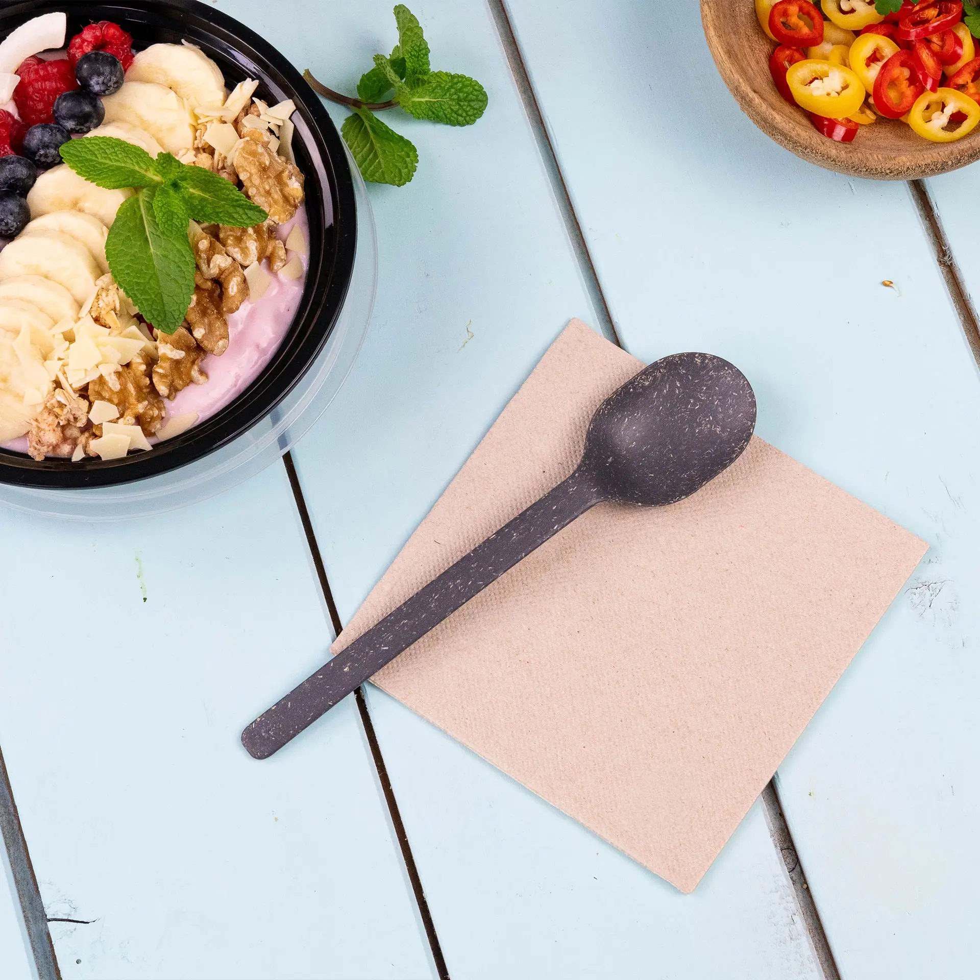 Bild eines schwarzen Löffels auf einer beigen Serviette neben einer Schüssel Joghurt mit Früchten und Nüssen auf einer blauen Holzoberfläche.